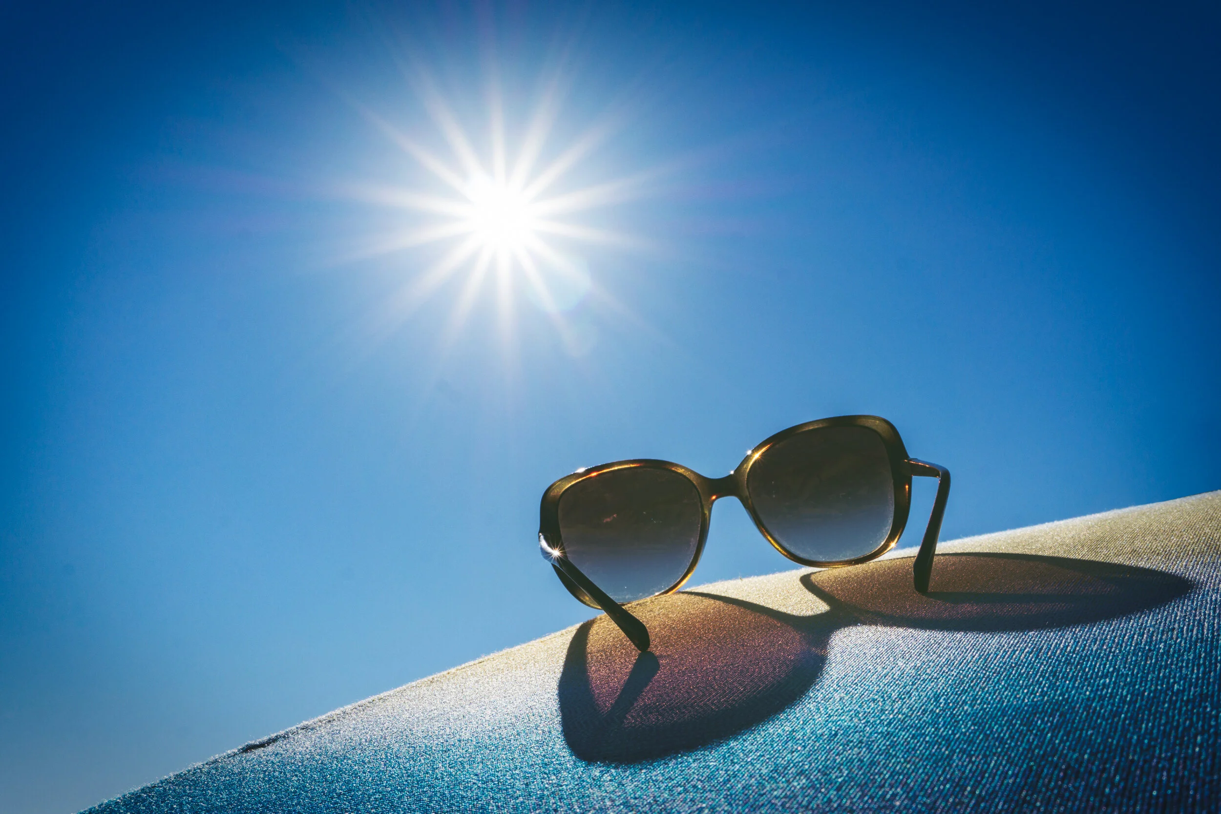 Sunglasses placed on a textured surface with the sun shining brightly in a clear blue sky