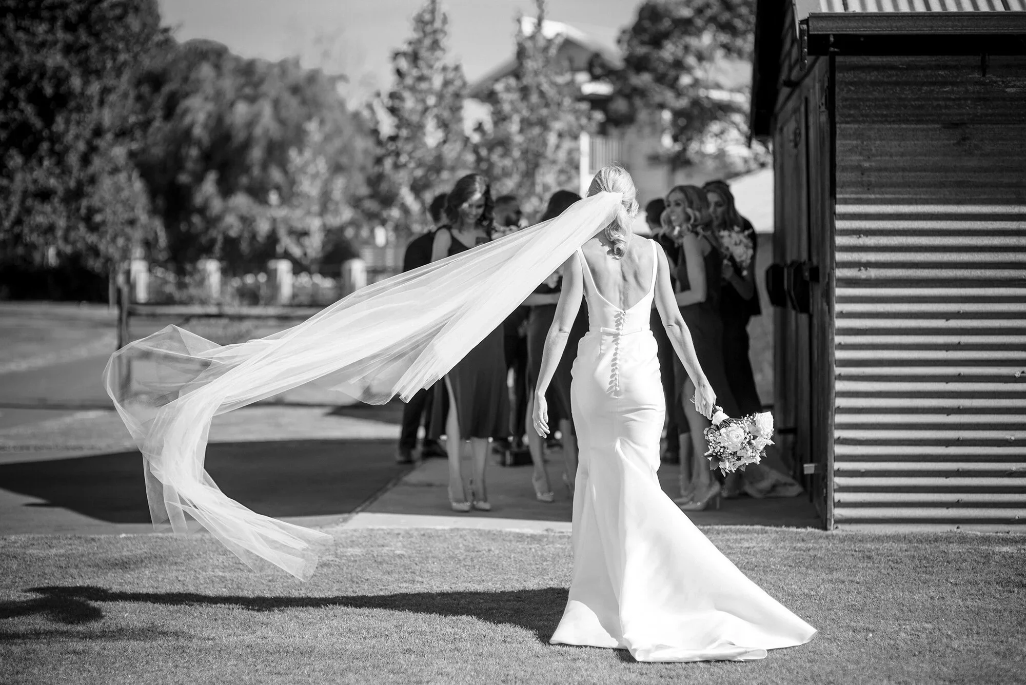 A bride in a fitted wedding dress holding a bouquet, walking out of a barn, with her veil flowing behind her and several women in dresses in the background.