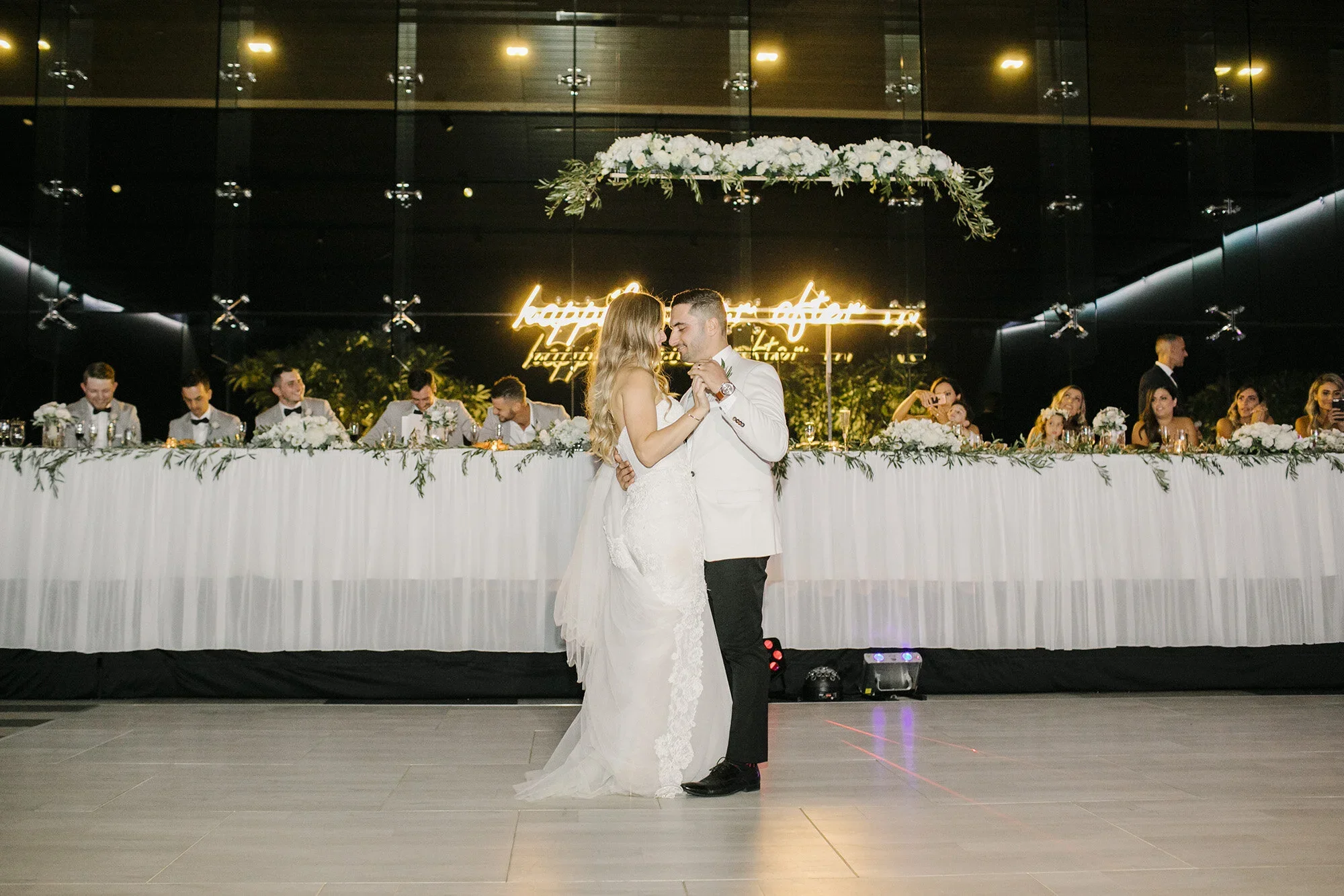 A bride and groom share their first dance at their wedding reception, with a long head table behind them decorated with white flowers and greenery. There is a neon sign with yellow writing in the background, and guests seated at the head table watching the couple dance.