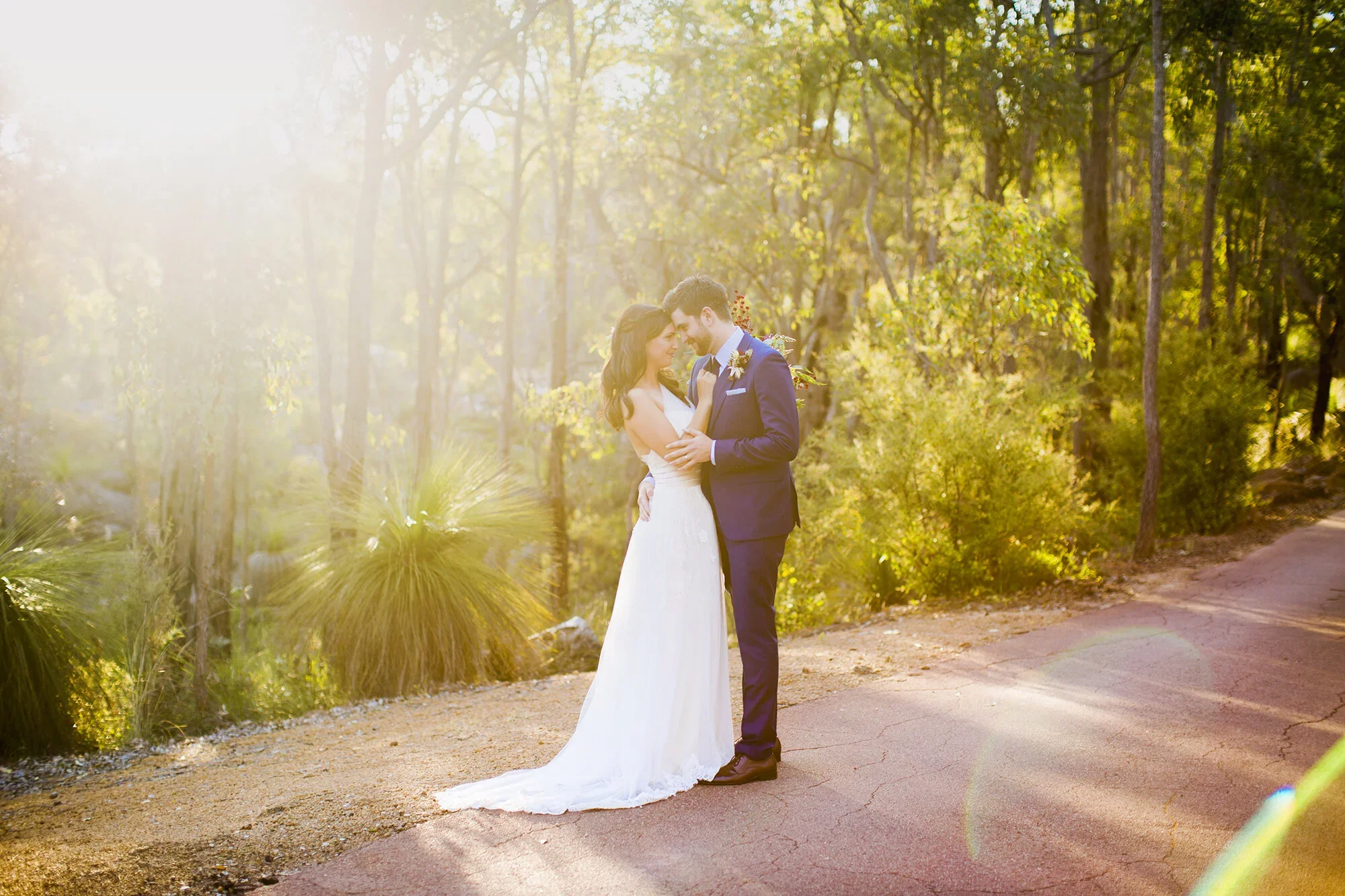 A bride and groom standing close together on a dirt path in a forest, smiling and embracing in wedding attire with sunlight filtering through trees.