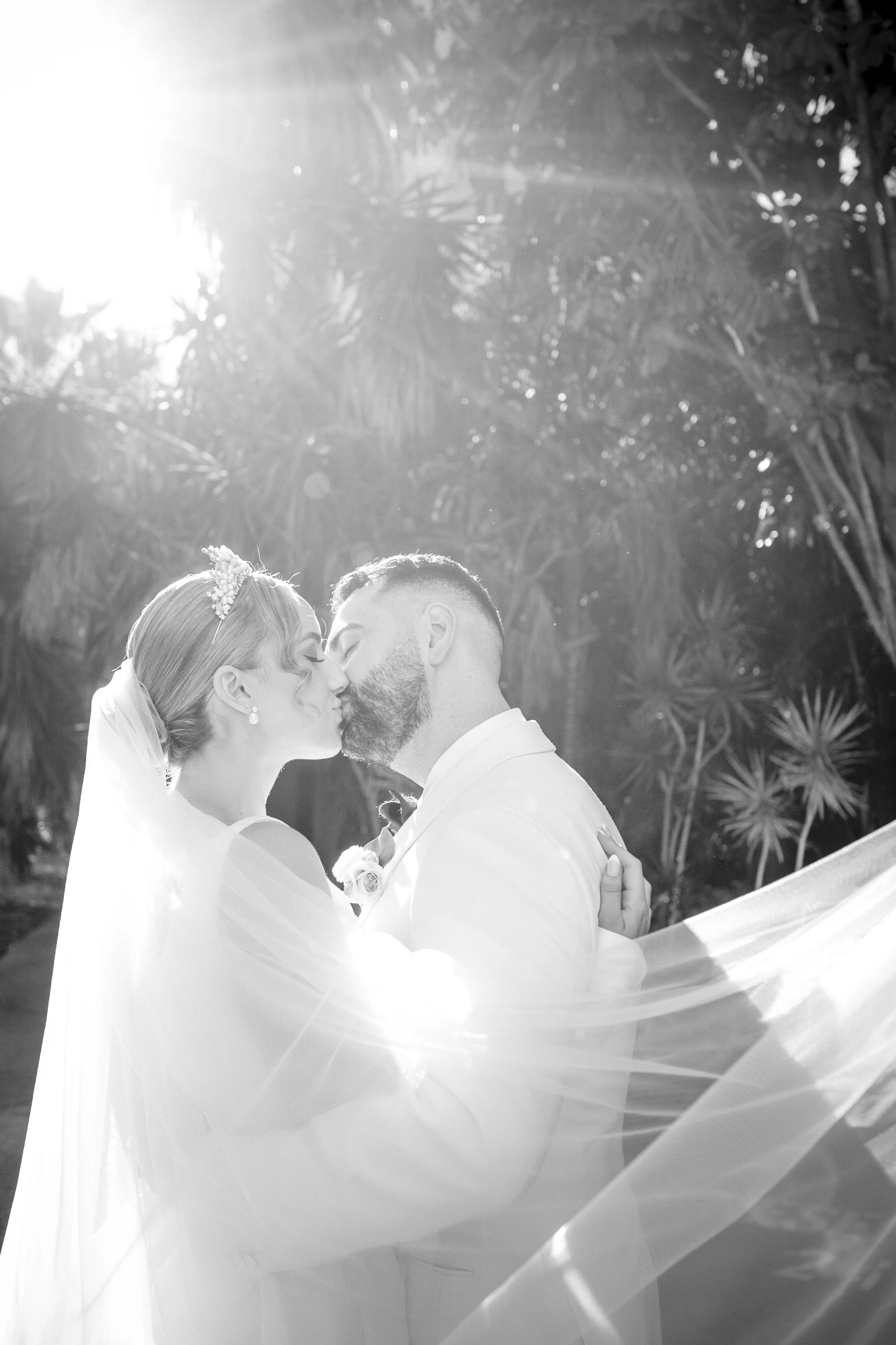 A bride and groom sharing a kiss outdoors, with sunlight filtering through trees in the background, in black and white.
