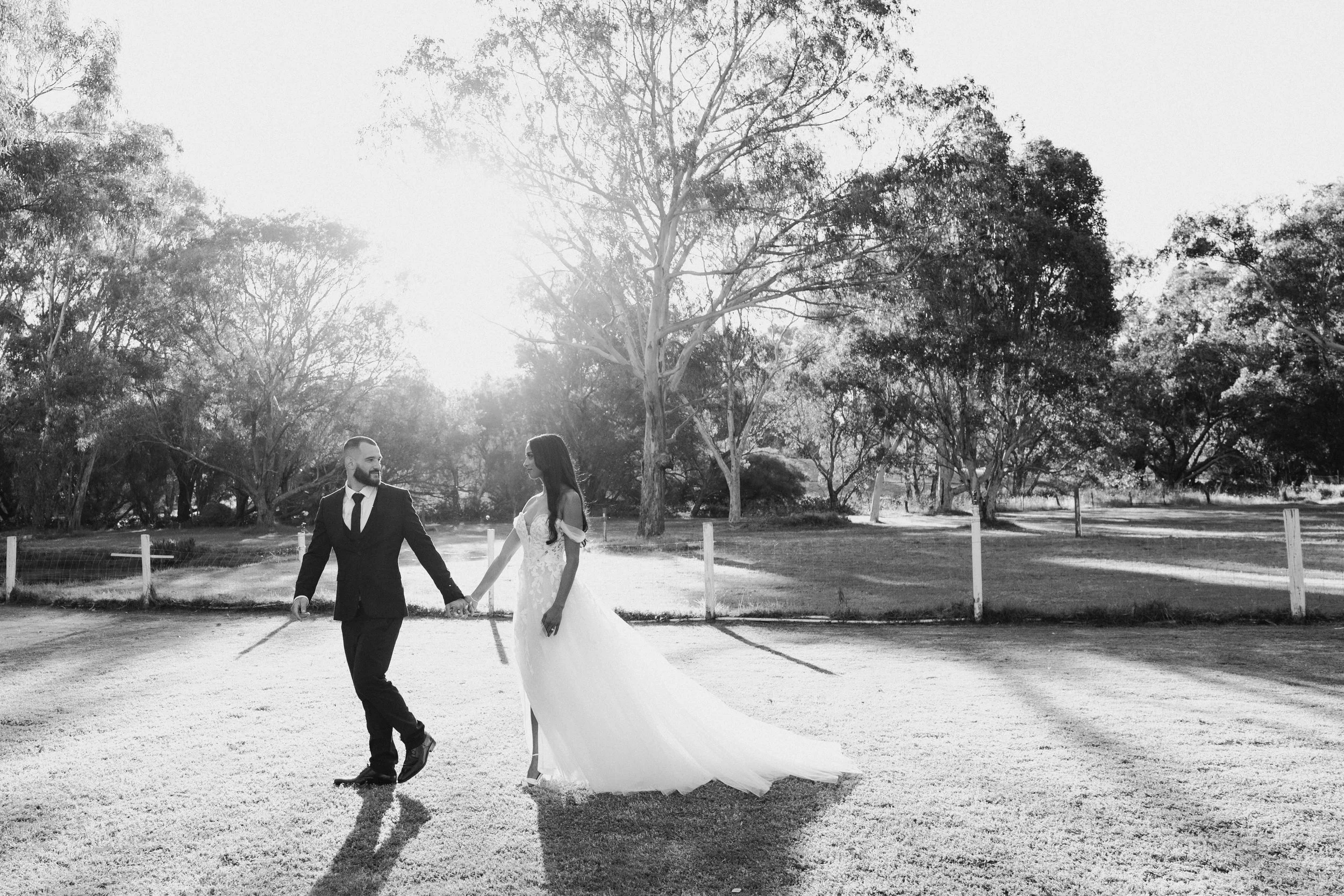 A black and white photo of a bride and groom holding hands in an outdoor park during sunset, with trees in the background.
