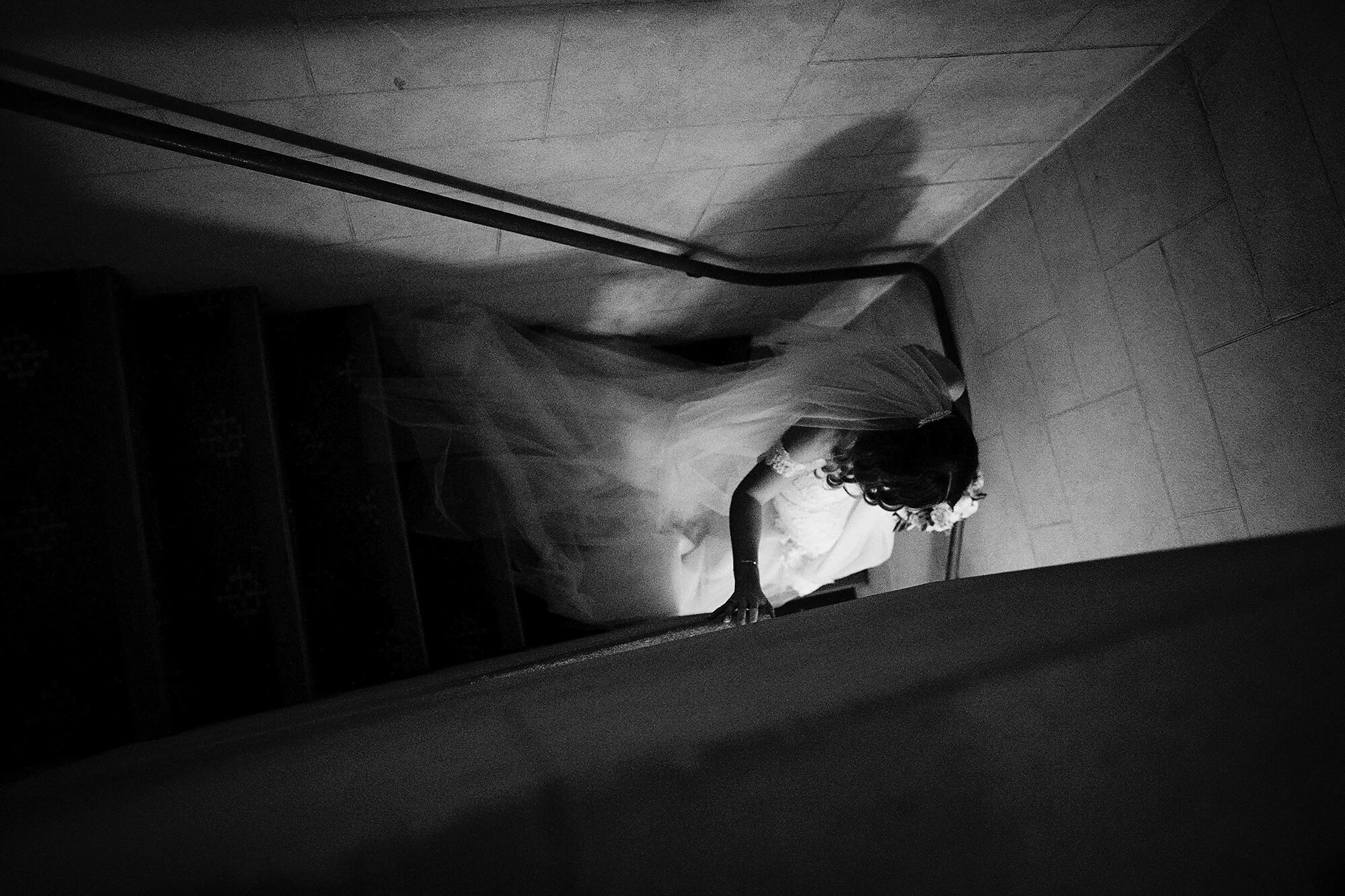 A young girl in a wedding dress, wearing a flower crown, looking down as she climbs a staircase in a dimly lit area.
