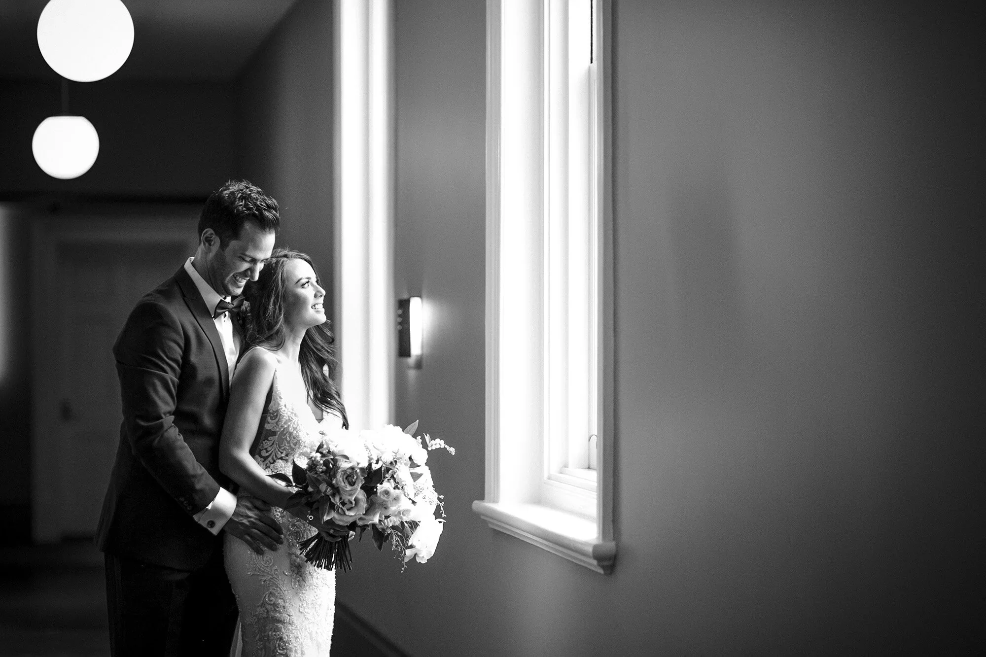 A black and white photo of a smiling bride and groom standing near a window, with the groom embracing the bride and the bride holding a bouquet of flowers.