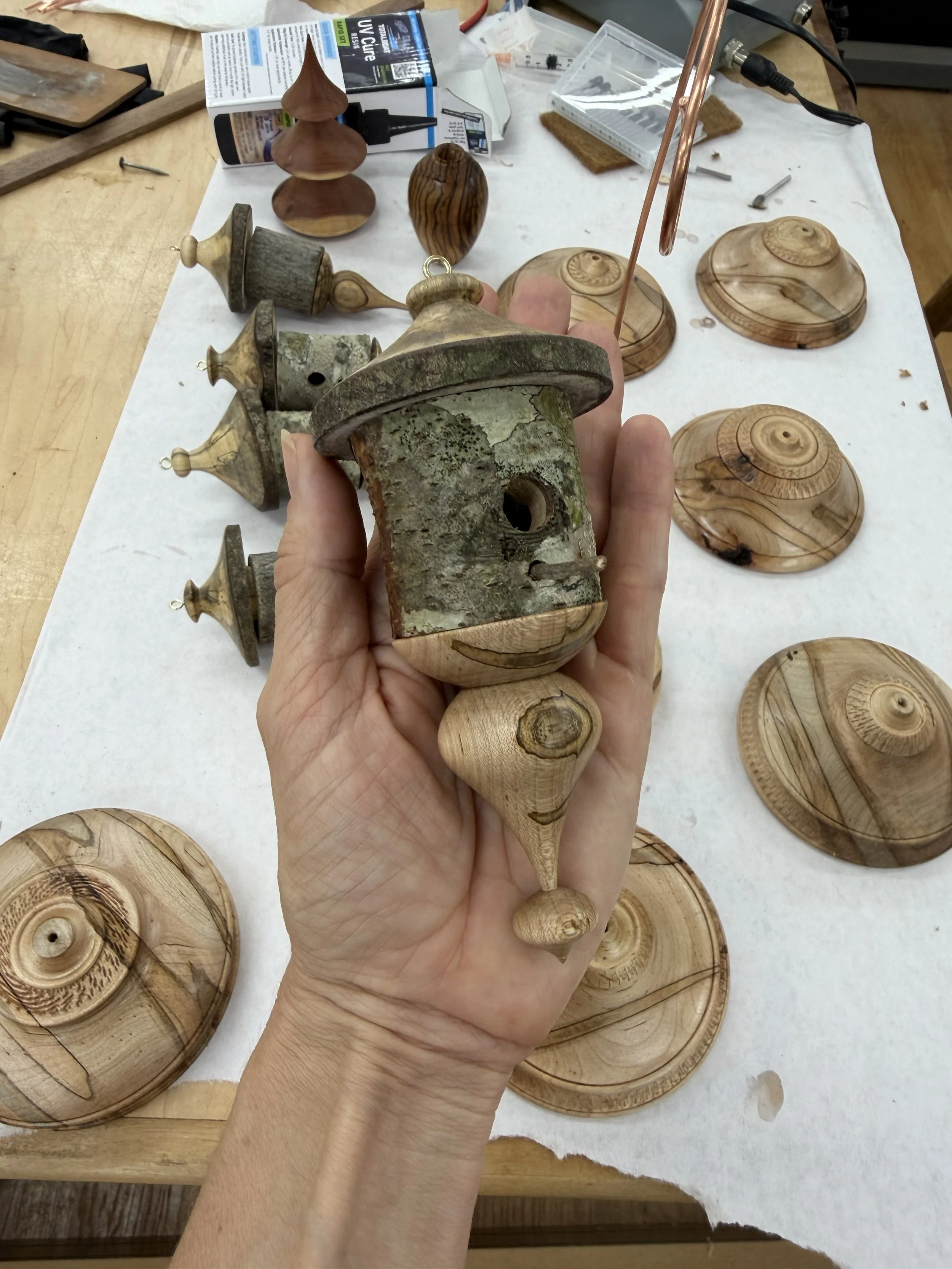 Hand holding a wooden birdhouse ornament over a workbench with turned bases and other birdhouse components awaiting assembly.