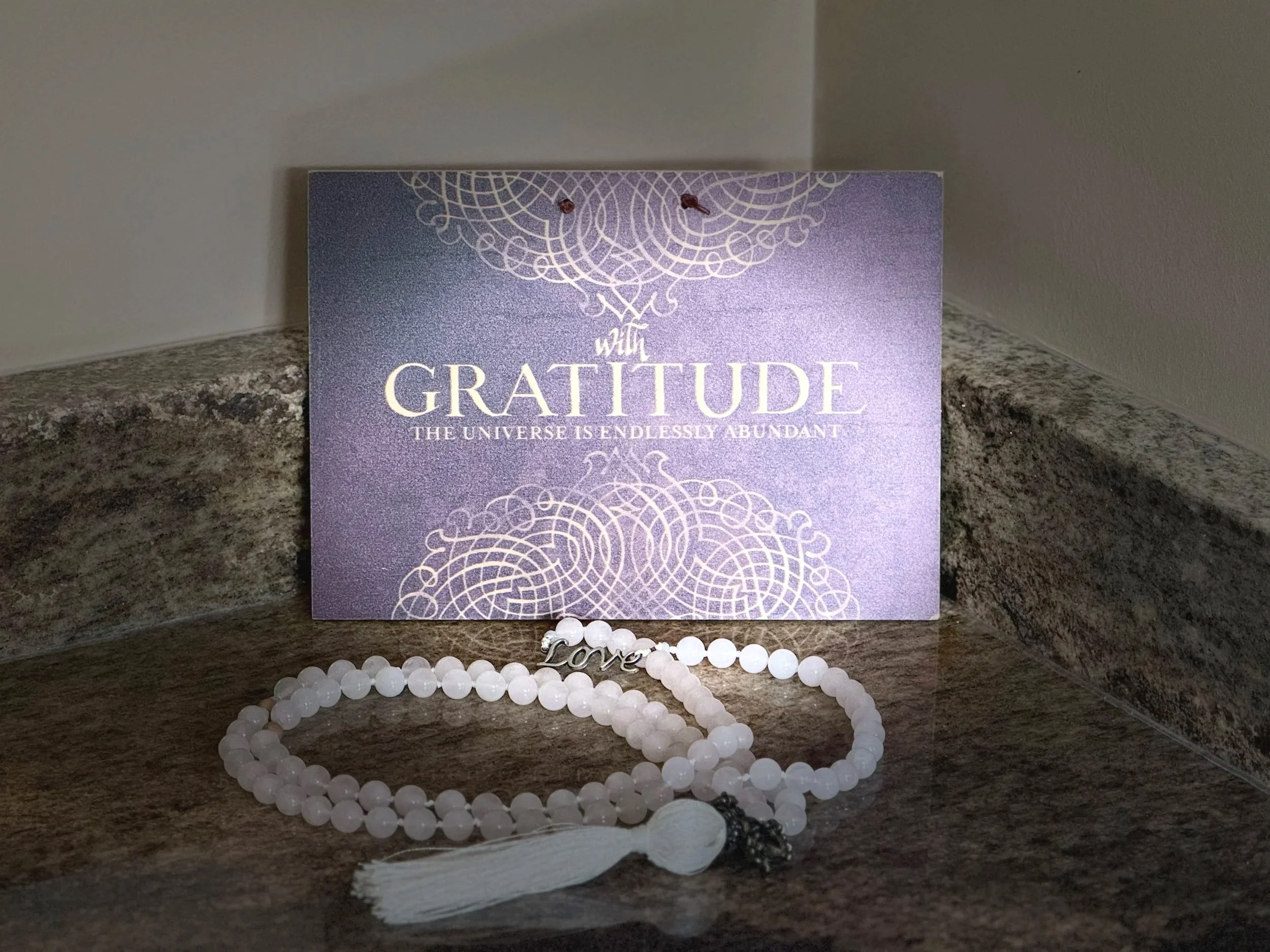 A plaque sits on the vanity. It reads, "With gratitude, the universe is endlessly abundant." In front of it is a mala strung with rose quartz beads. It has a metal bead that says "love" and a white tassel.