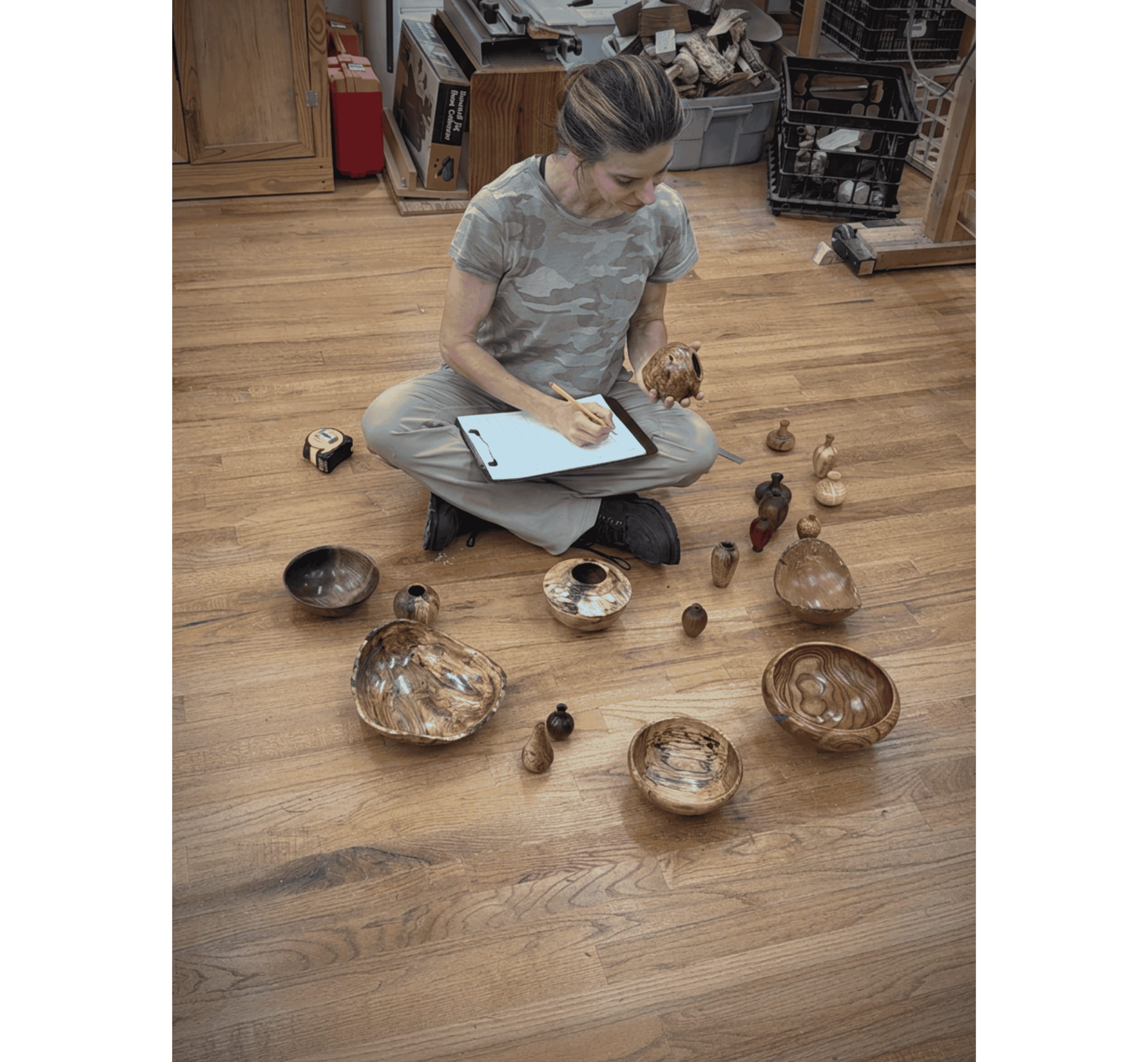 Rebekah sits on the floor of her woodworking shop, surrounded by her turnings. She has a clipboard to record her inventory in her lap, and is taking notes with one hand while holding a hollow form in her other hand.