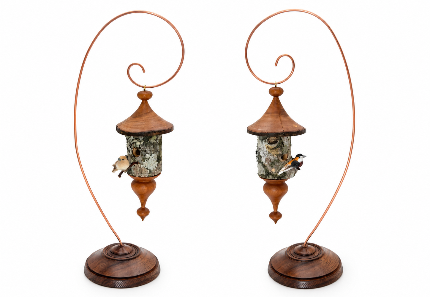 Two handcrafted wooden birdhouse ornaments displayed side by side on curved copper-wire stands with turned wooden bases. Each features a natural bark body, turned wooden roof and finial, and a tiny decorative bird.
