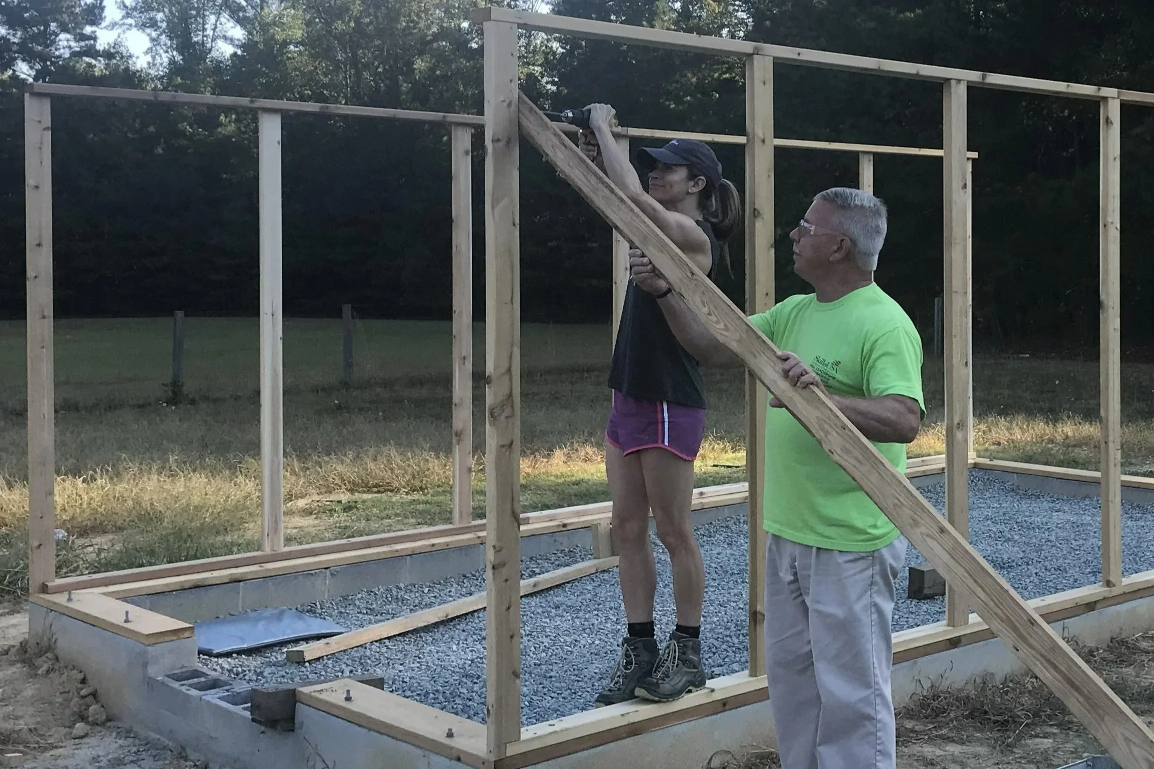 Rebekah and Mr. Pendergrast work on framing the walls of the greenhouse.  Mr. Pendergrast holds a piece of wood for bracing the wall while Rebekah uses an electric drill to install a screw to hold it in place.