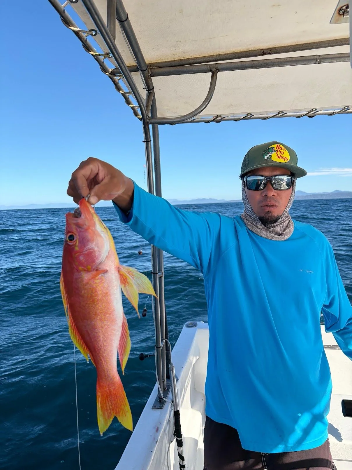 Man holding a pink and yellow fish on a boat out at sea, wearing sunglasses, a green hat with a fishing logo, and a blue long-sleeve shirt.
