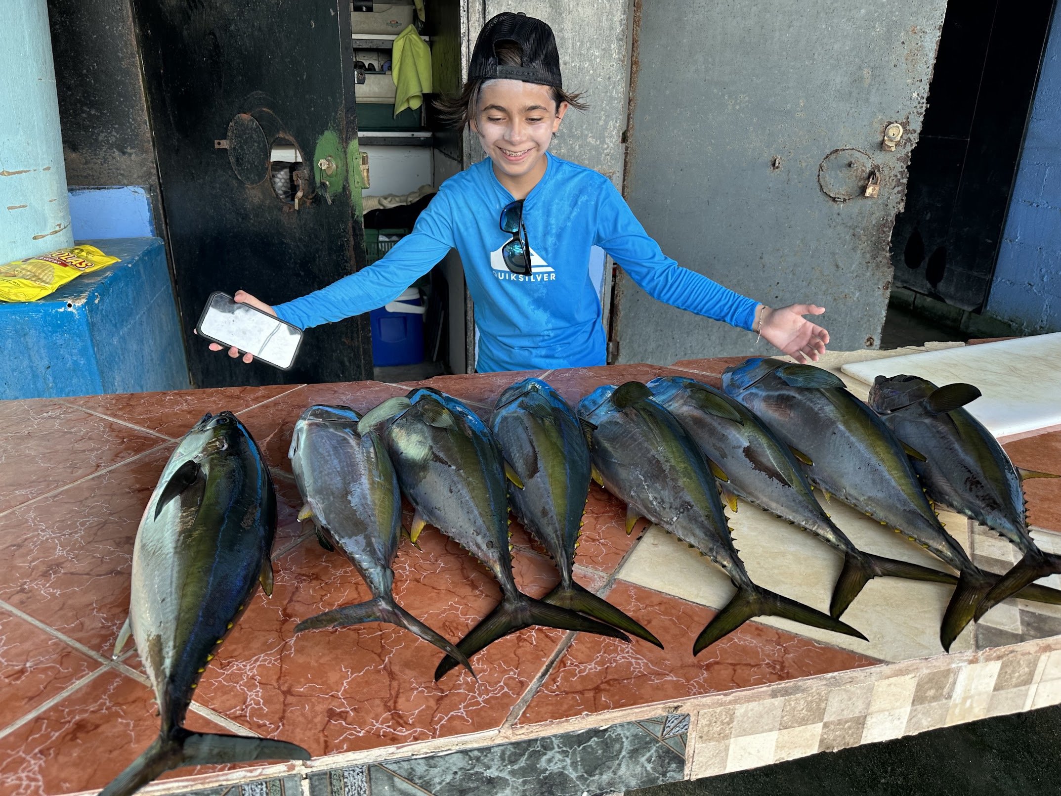A young boy wearing a black cap and a blue Quicksilver shirt stands behind a table with freshly caught fish arranged in a row. He is smiling and holding a smartphone in his right hand, with sunglasses hanging from his shirt collar. The background fea