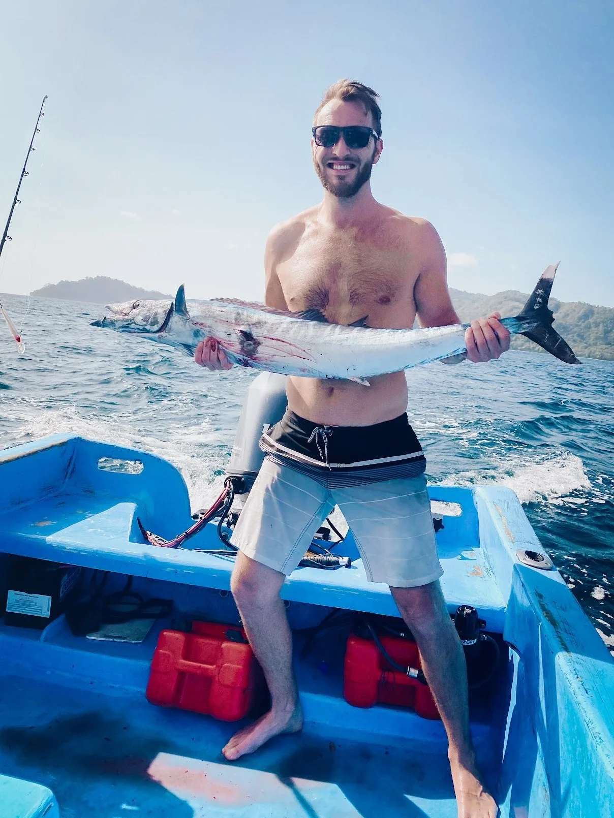 A man with sunglasses and a beard, shirtless, holding a large fish on a boat in open water with islands in the background.