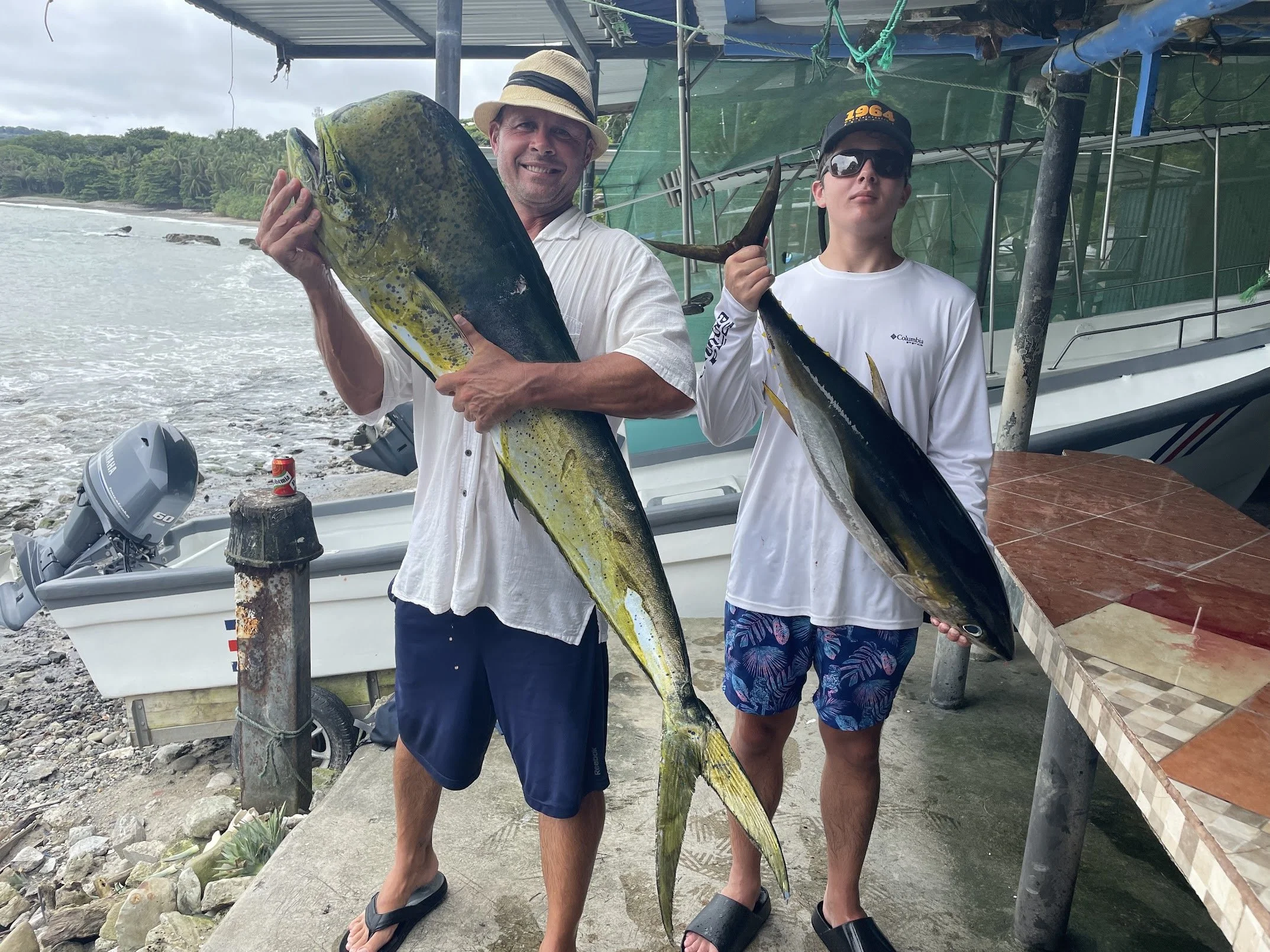 Two men standing on a dock holding large fish, with a boat and shoreline with greenery in the background.