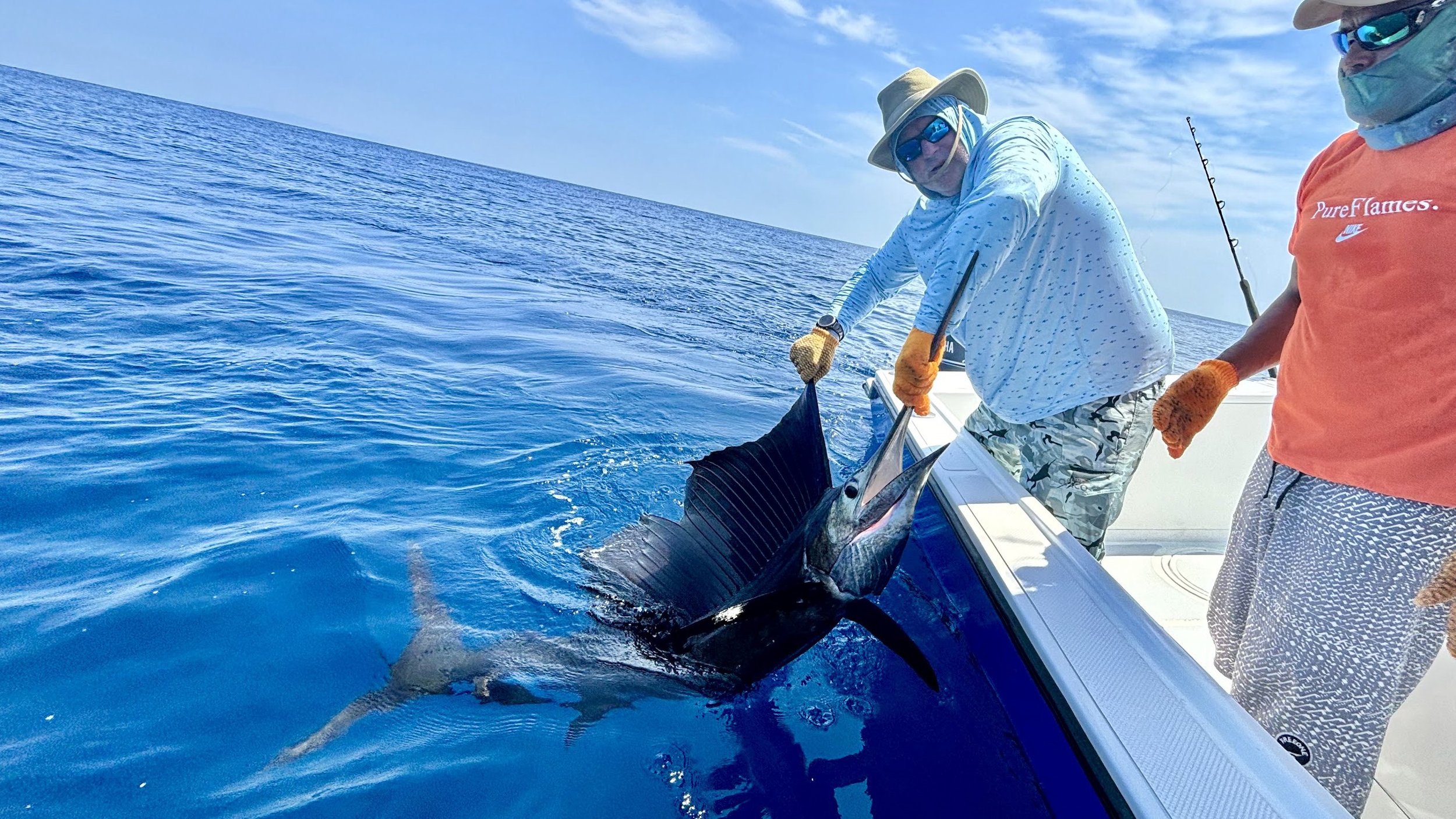 Two men on a boat catching a large fish in the open ocean, one holding the fish and the other assisting.