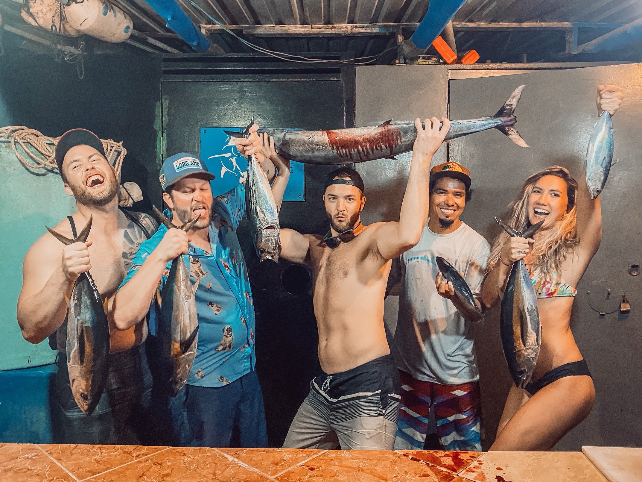 Group of five people, four men and one woman, holding large fish, posing in an indoor setting with a dark wall and fishing gear in the background.