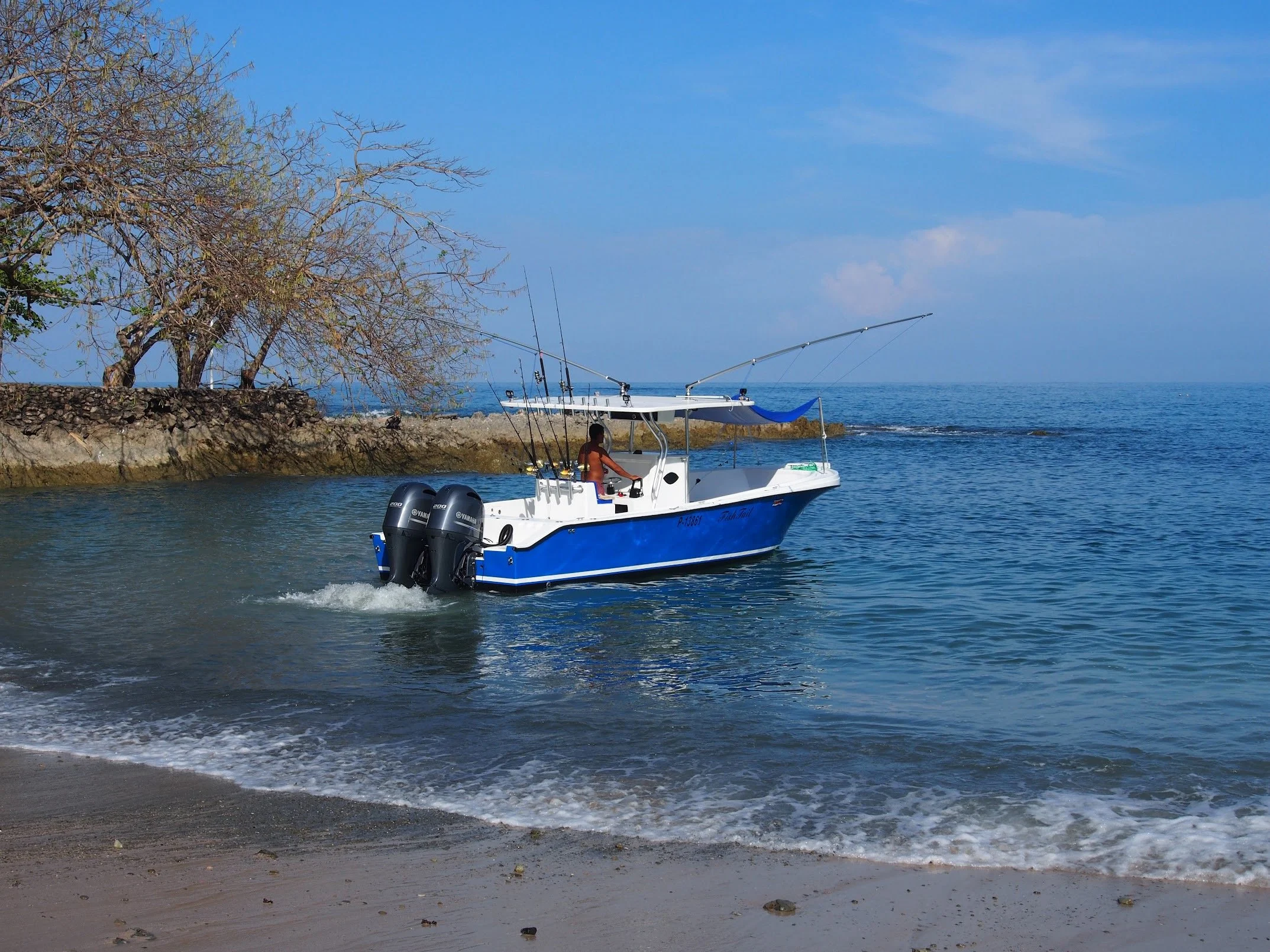 A boat with two outboard motors sailing past a rocky shoreline with dry trees under a blue sky. Fishing Boat Tour Charter