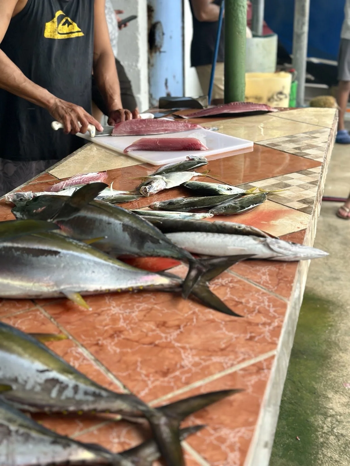 A person is preparing fresh fish at a market stall, with various fish and filleted fish on a tiled table.
