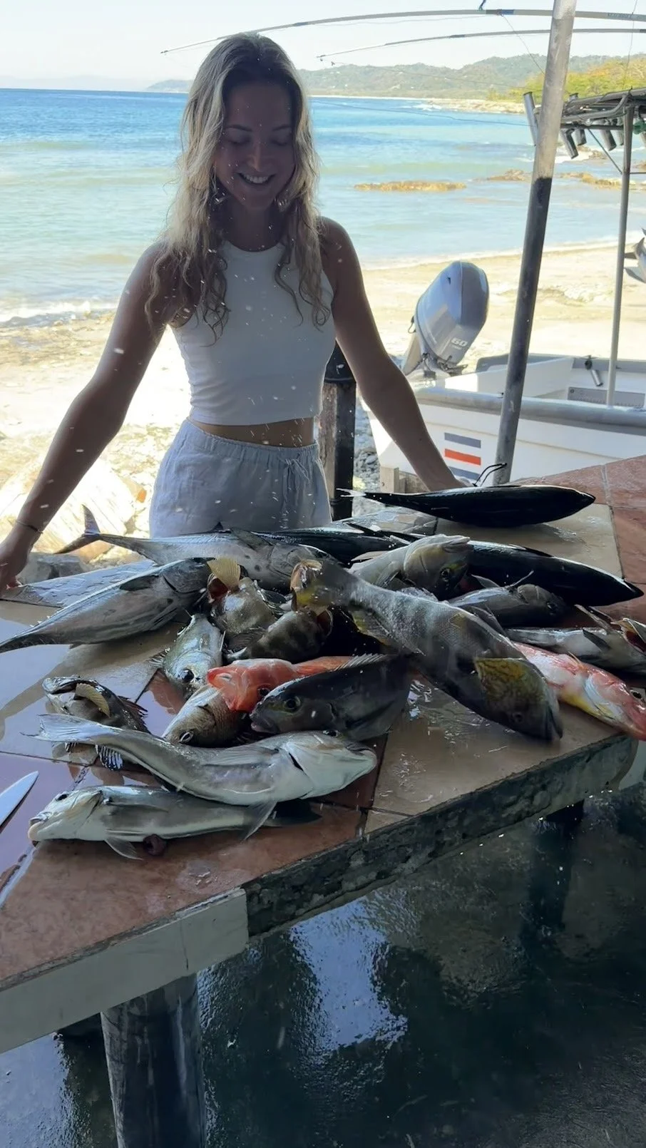 A woman in a white crop top and shorts standing behind a table full of freshly caught fish at a seaside location with boats and water in the background.