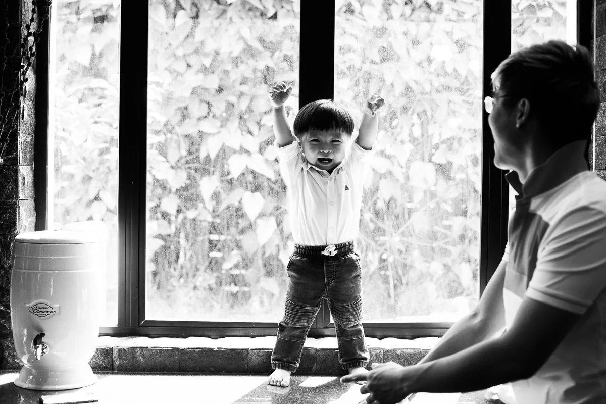 A little boy stands before a window with his arms raised up above his head in victory.
