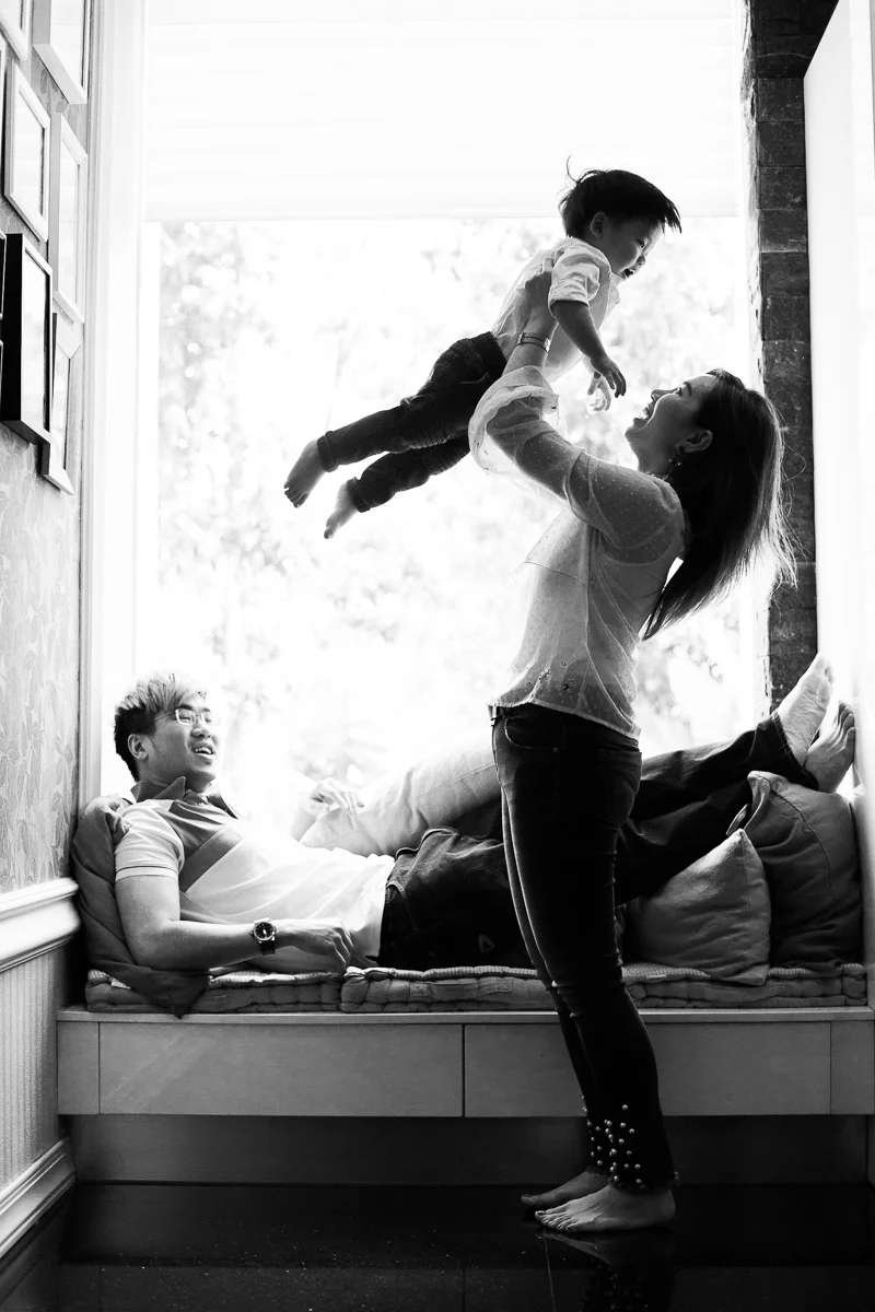 A mother plays with her toddler son by lifting him overhead while a father looks on and smiles by family photographer Erica Knecht.