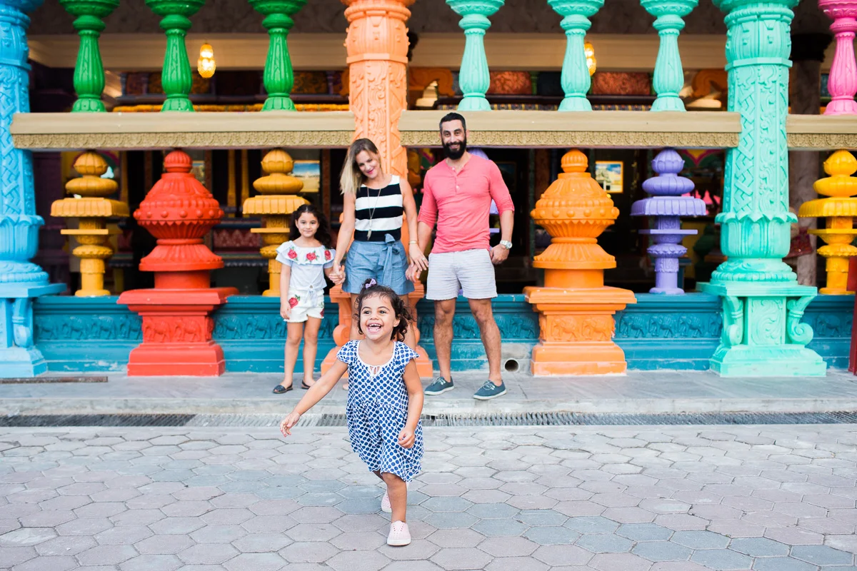 A family stands together as their little girl runs towards the camera with an exuberant smile on her face at Batu Caves.