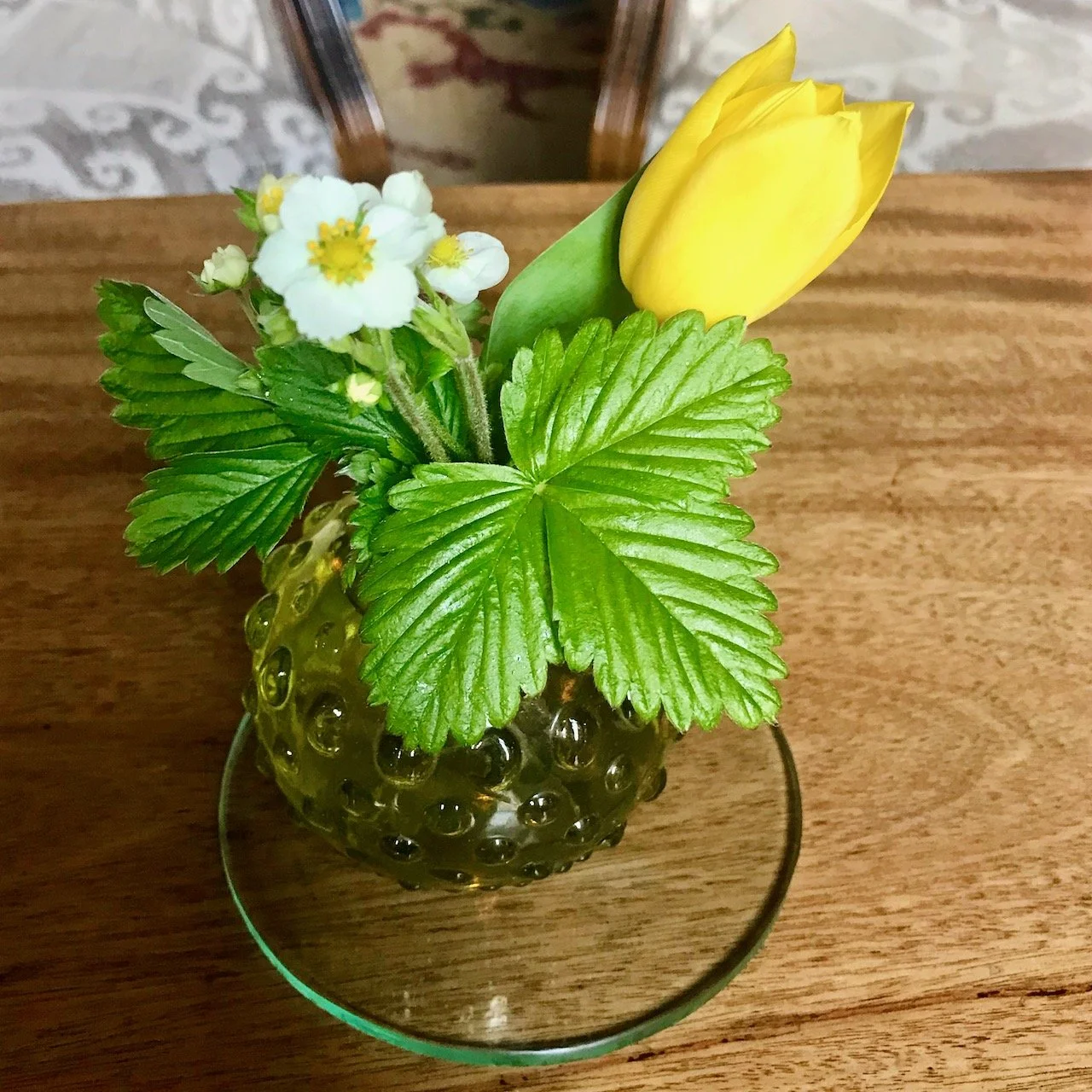 A yellow tulip and strawberry flower and leaf in a small glass vase