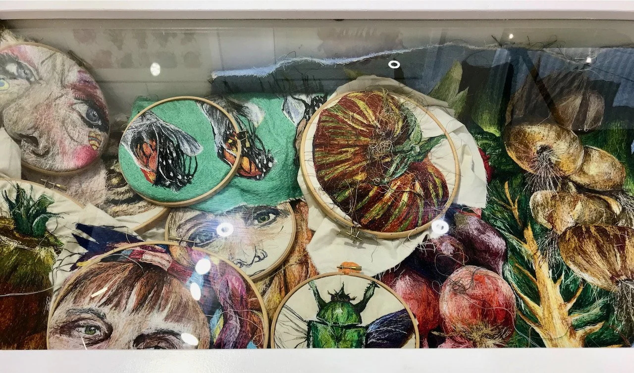 Looking into a glass display case onto many embroidery hoops, including some of vegetables, bugs and portraits