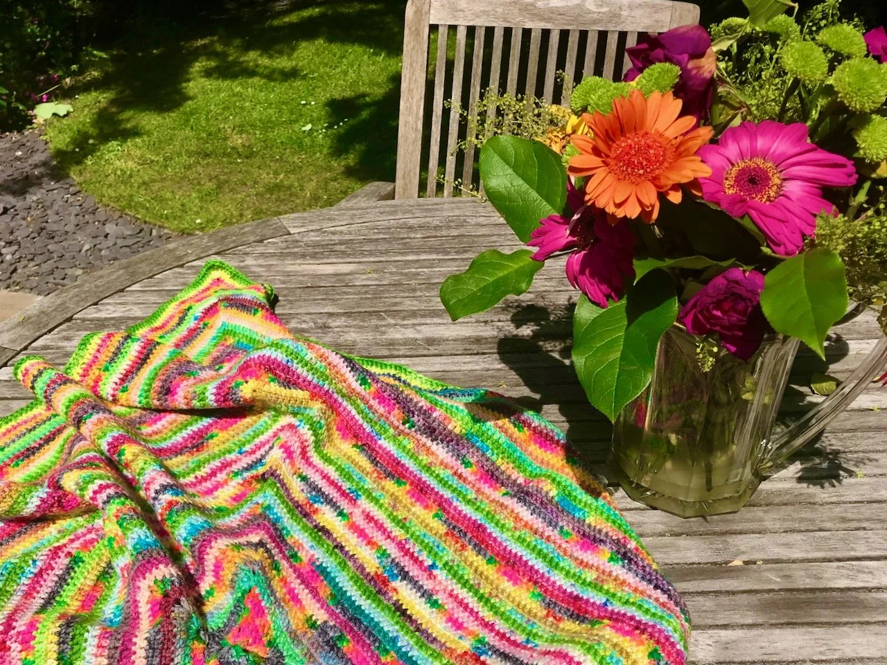 A brightly coloured crochet throw on a garden talbe with a vase of brightly
