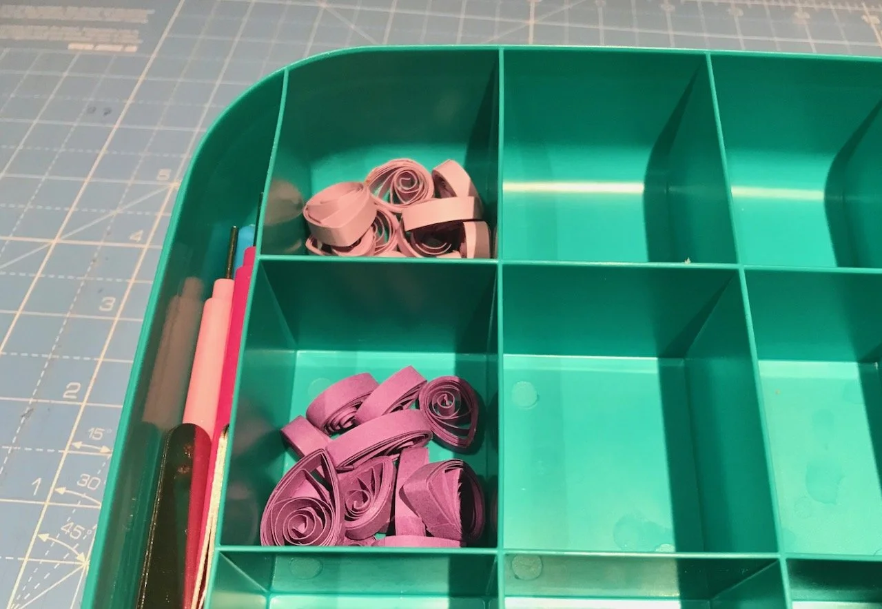 A green plastic tray with segments containing lilac and purple quilled paper, and a longer section containing the quilling tools