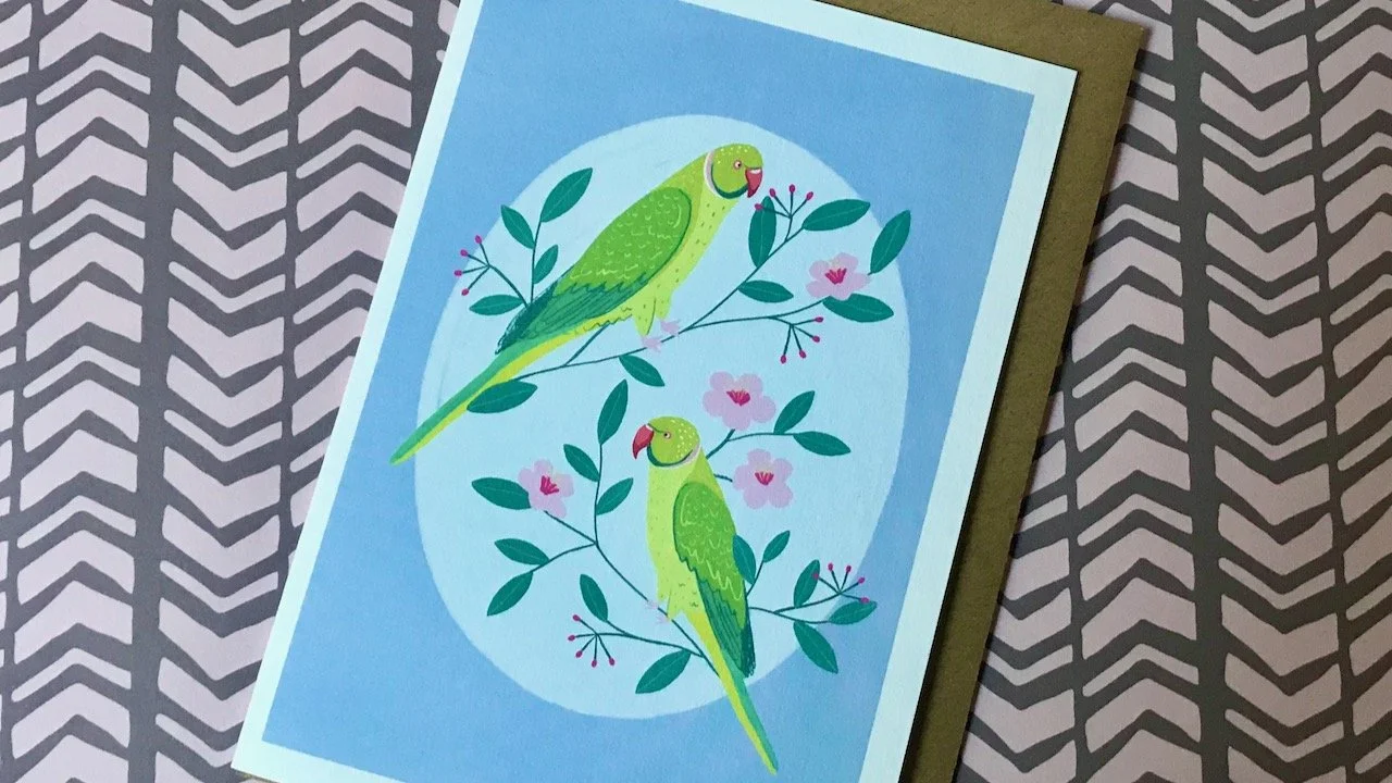 Two green budgerigars sitting on branches against a light blue oval on a darker blue background