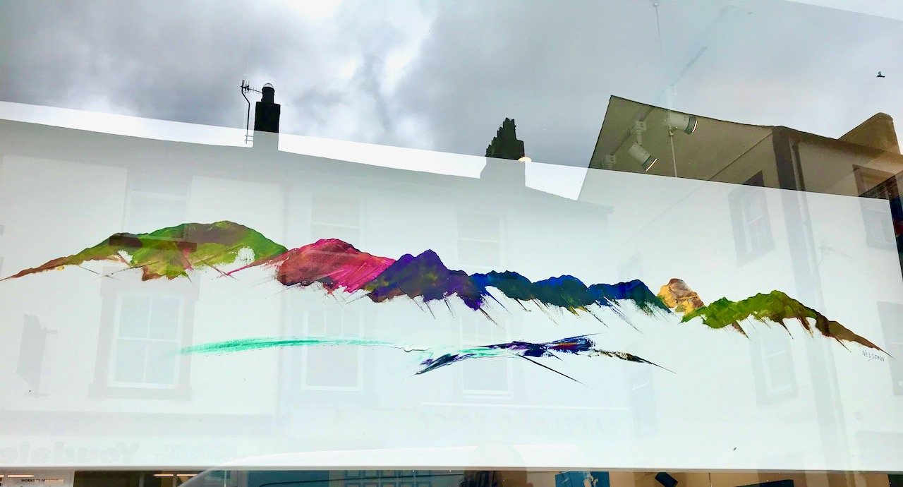 A painting of mountains in a Lake District shop window with the town in the reflection