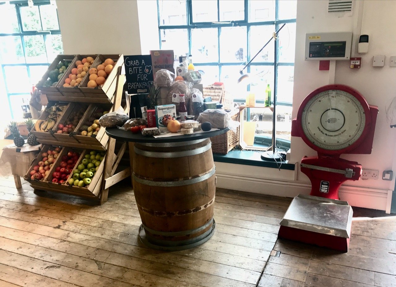 In the farmshop with a display of fruit in crates on the left, a large barrel table displaying groceries and on the right next to the door a red old fashioned (large) weighing machine
