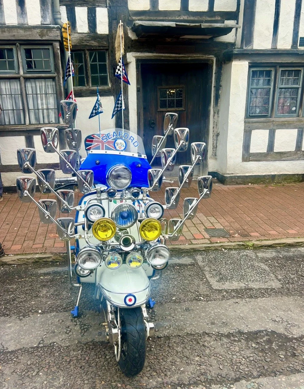 A scooter with plenty of wing mirrors and headlamps, a flag or two and parked outside a half timbered building