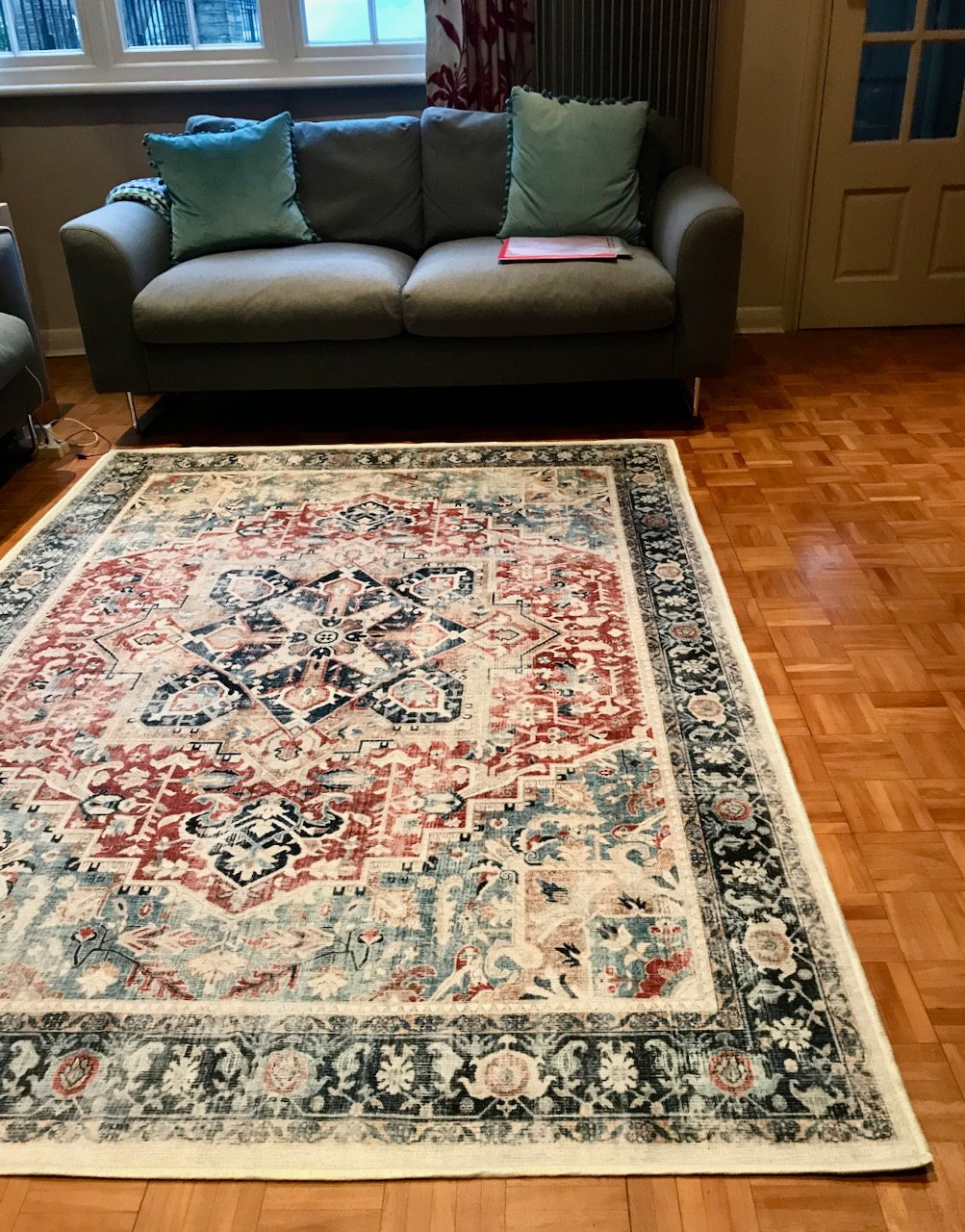 the new rug in the foreground on a wooden floor, with a grey two seater sofa at the rear