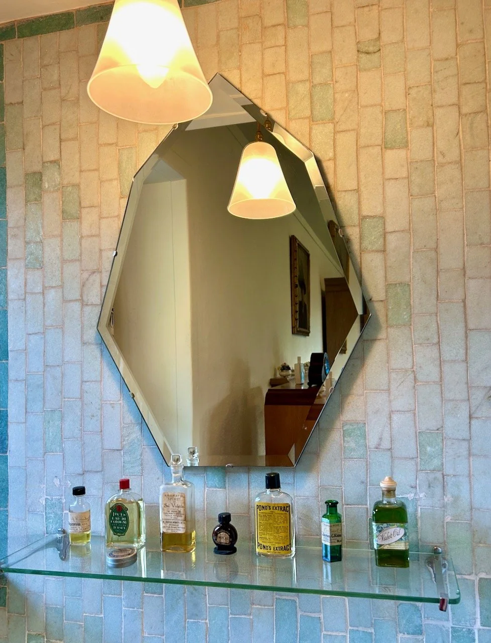 Delicate coloured blue tiles in the bathroom with a shaped bevelled mirror and various bottles on a glass shelf