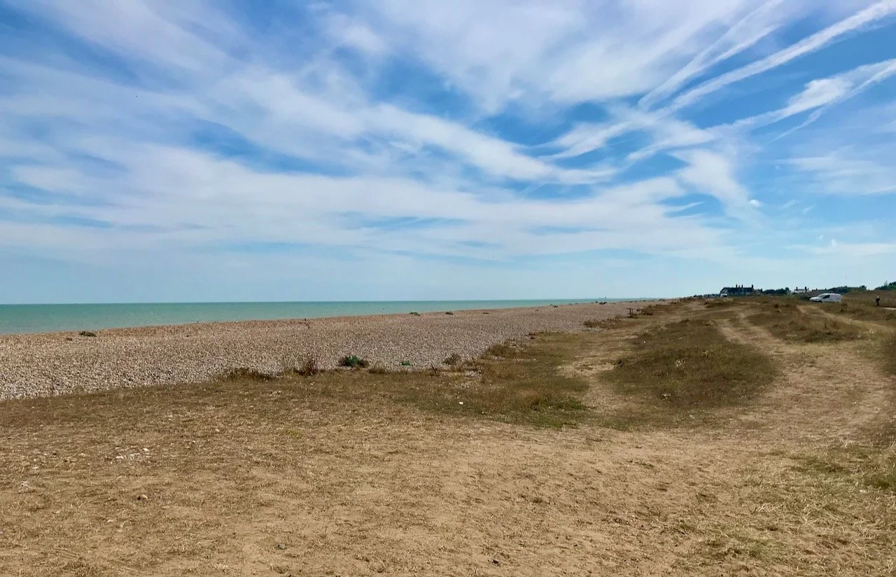 looking towards the sea over the scrub land and stony beach - a blue sky with unusual clouds above