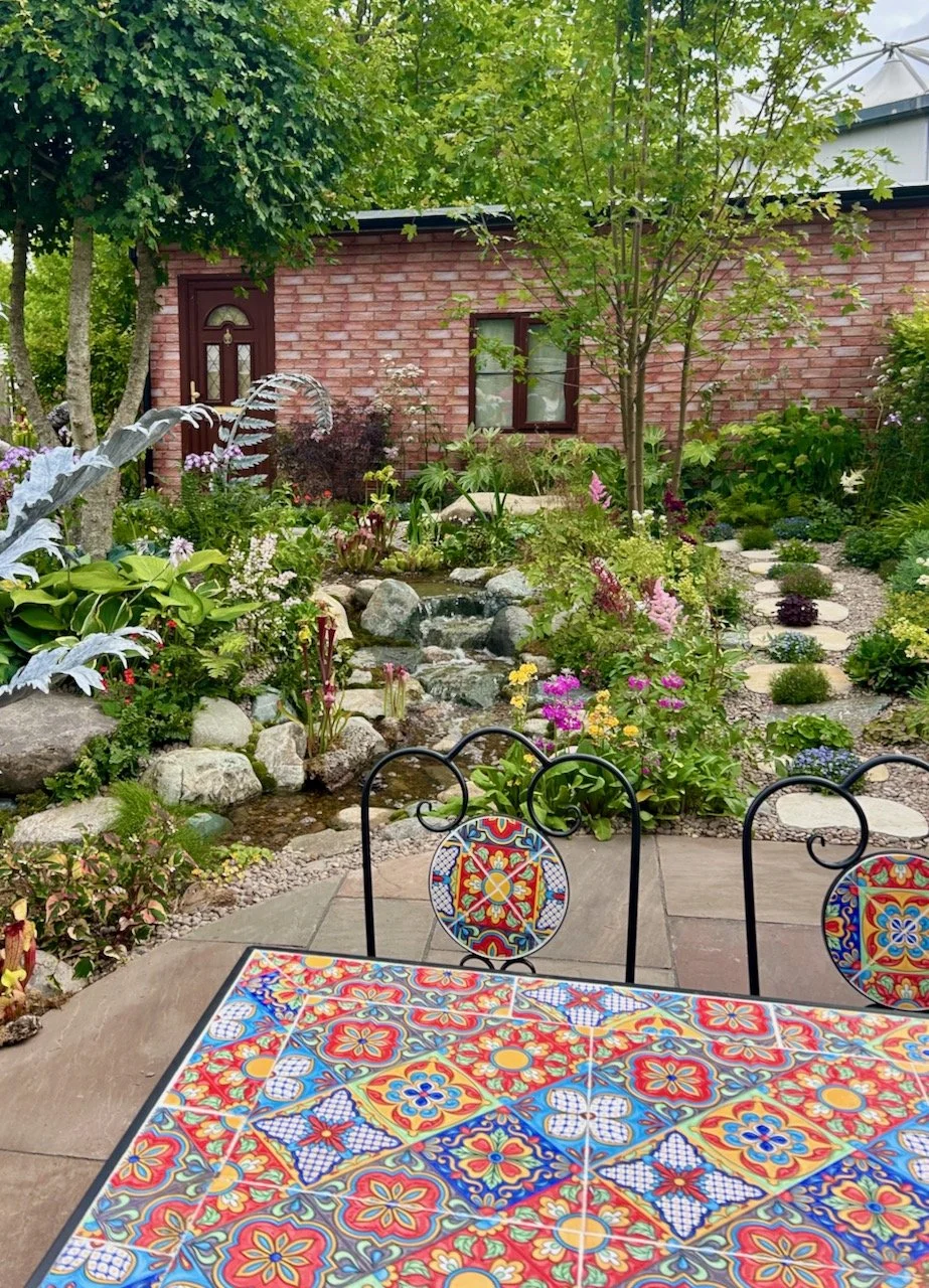 The foreground shows a brightly coloured mosaic table and chairs on a paved area, on the left is the gunnera shaped waterfall (just) with more trees which will screen the house when grown