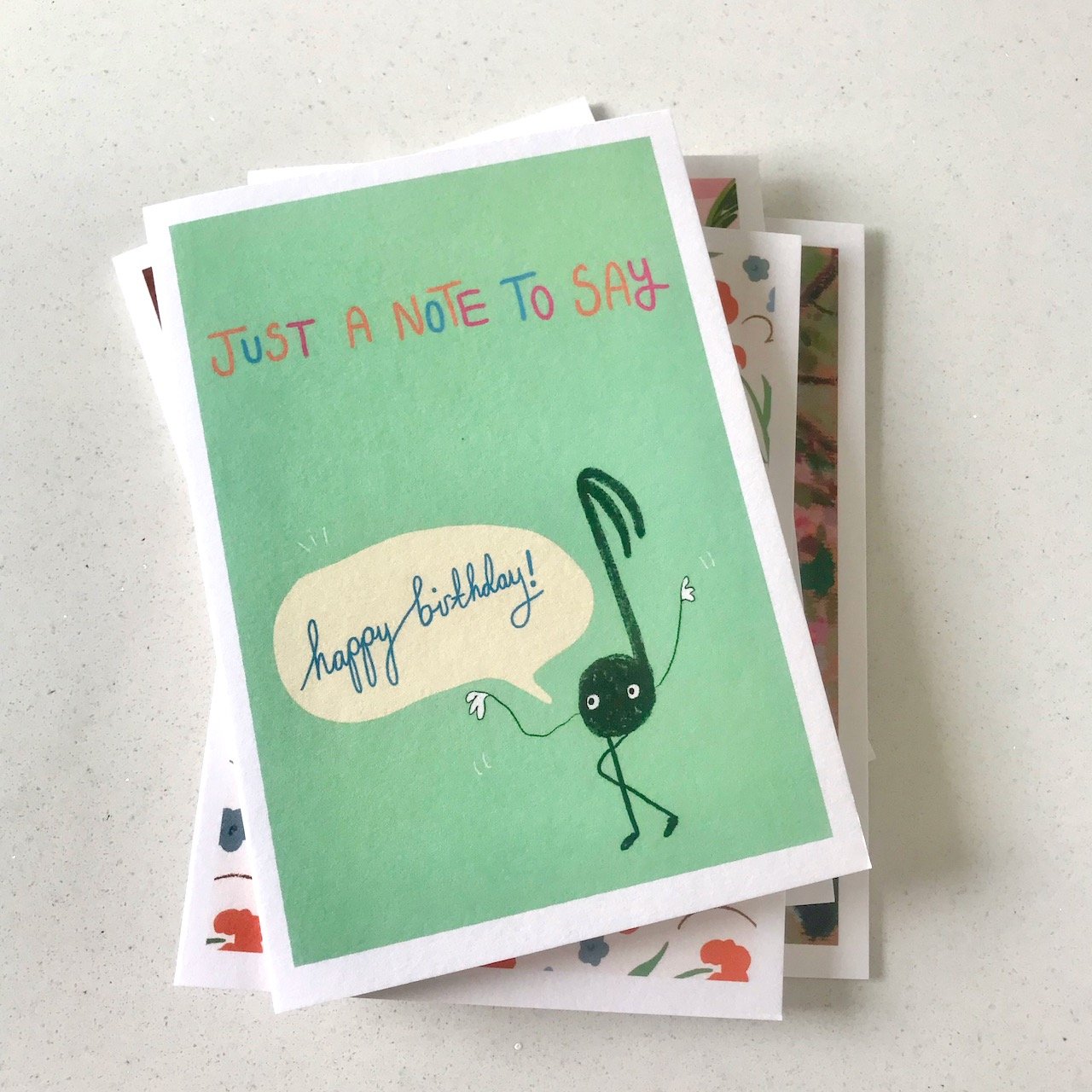 A black musical note with arms and legs on a green background with a speech bubble for 'happy birthday' above the illustration is the text Just a note to say written in orange, pink and blue on a pea green background