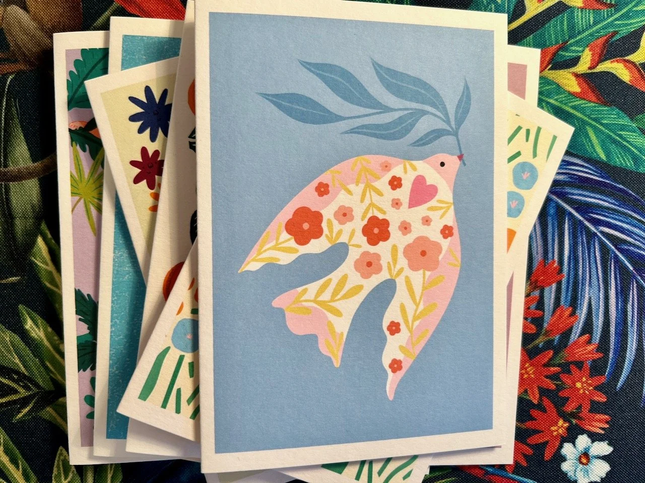 A pink and white dove with a floral pattern carrying an olive branch on a pale blue background