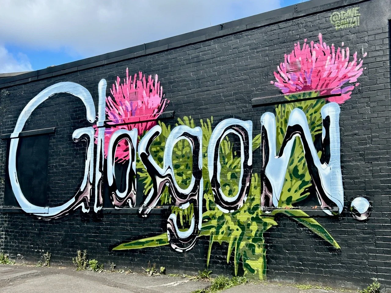 Glasgow street art - the town's name in white on a black brick wall with two thistles in pink and green leaves behind