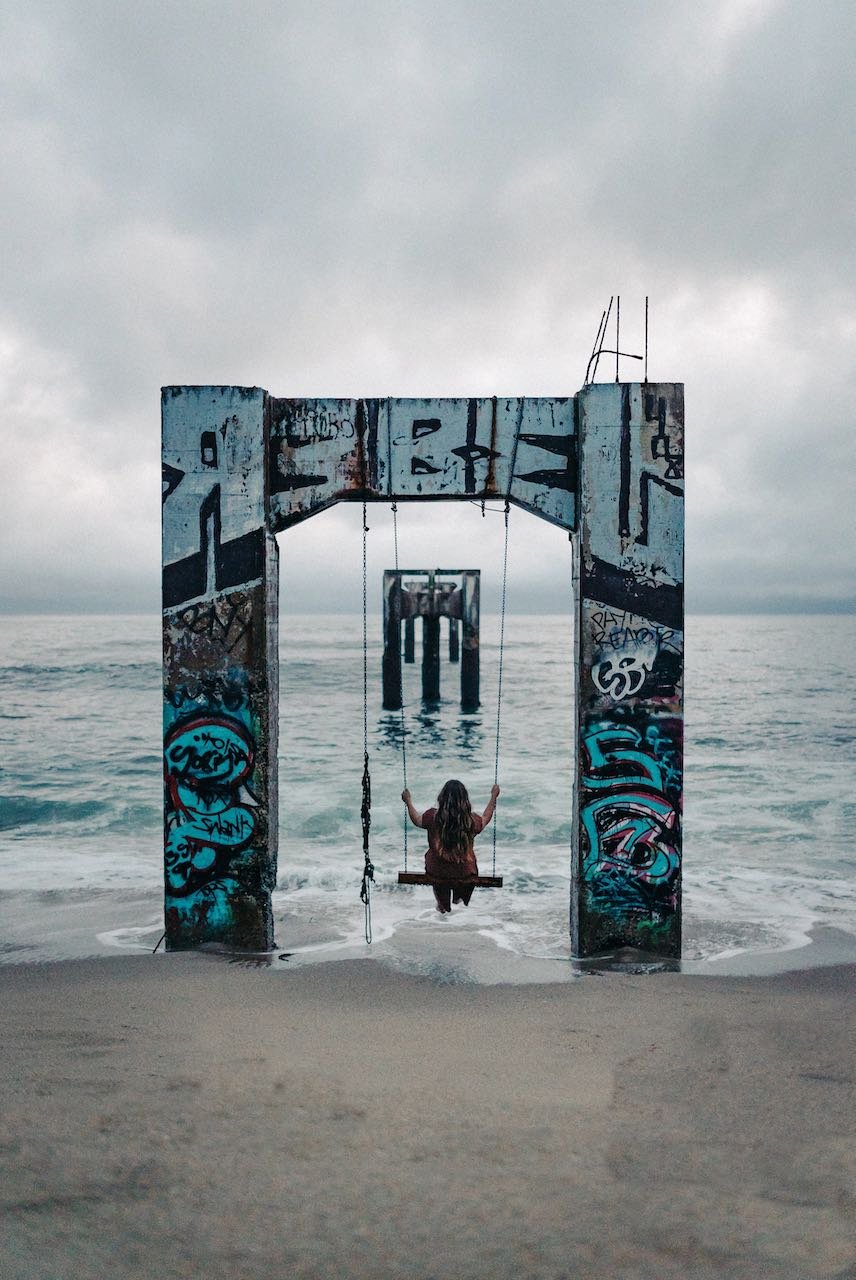 Woman on a swing made from big, graffitied concrete blocks, at the sea's edge with further concrete blocks into the distance