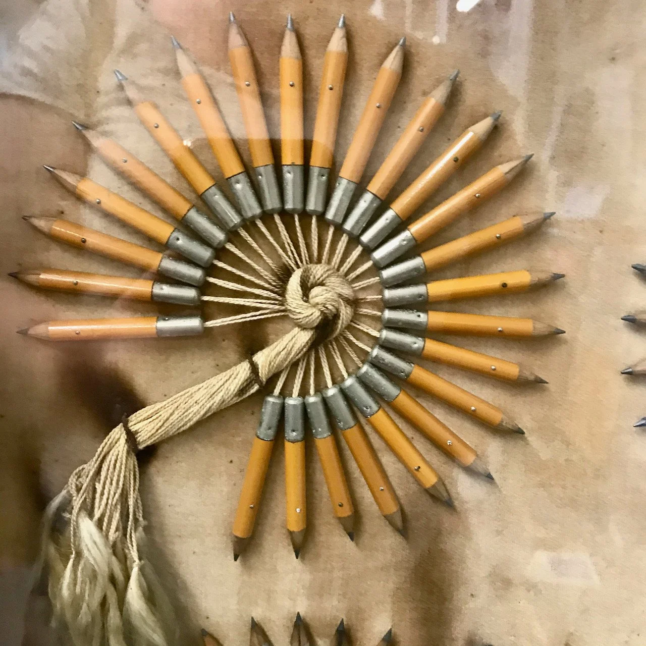 A close up of the same display showing short yellow pencils in a 3/4 circle (with the lead facing outwards) - in the inner circle the string from the top of each pencil is entwined and twisted to form a knot and 'tail' for the shape
