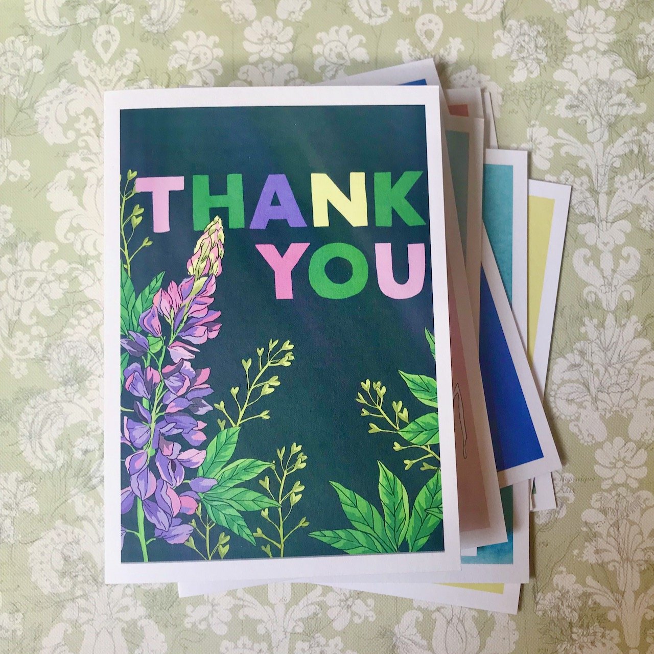 A navy blue background with capital letters in pink green purple and yellow spelling THANK YOU.  in the lower part of the card is a pink/purple lupin plant with green leaves
