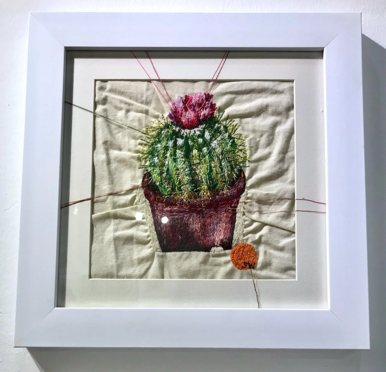 An embroidered potted cactus with a flower on top on crumpled cream fabric in a white frame.  Threads extend from the picture over the picture mount.