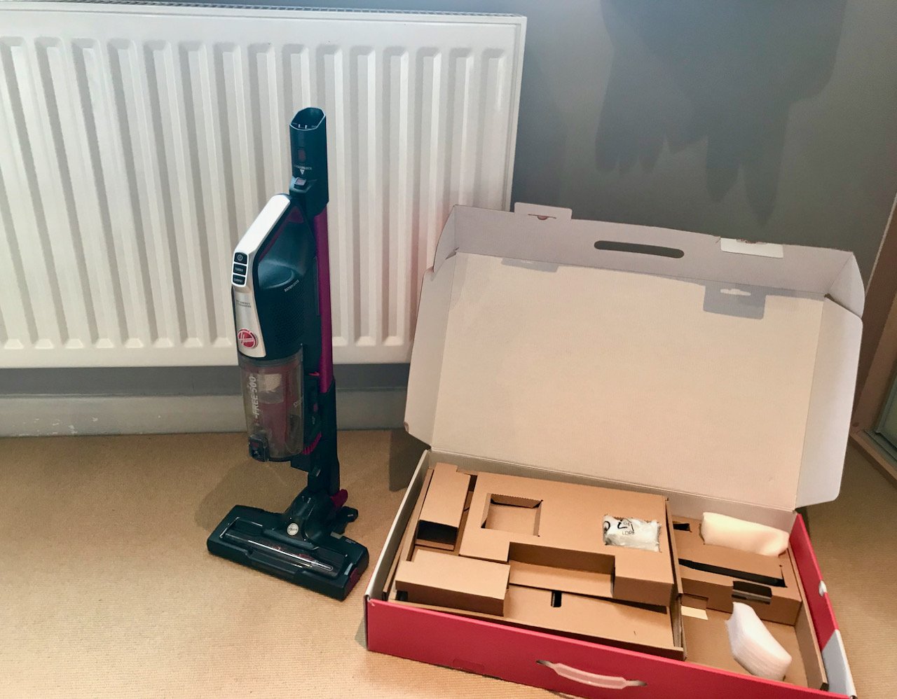 the cordless vacuum cleaner in compact mode – which is shown against the radiator and is approximately three quarters of the height of the radiator, the empty box with packing is alongside on the floor.