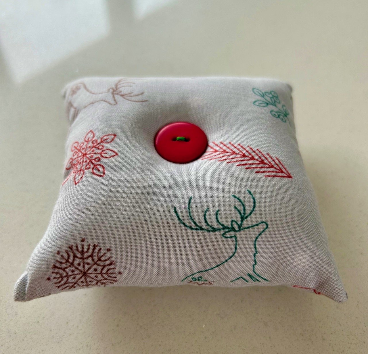 A light grey square fabric pincushion, the material has snowflakes arrows and reindeers in red, green and brown line drawings - and there's a large red button at the centre