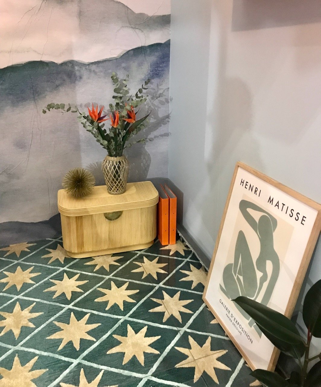 Below the open shelving unit showing the floor with a blue/green rug with stars on there is a small wicker-like storage unit and a framed matisse print leaning against the wall