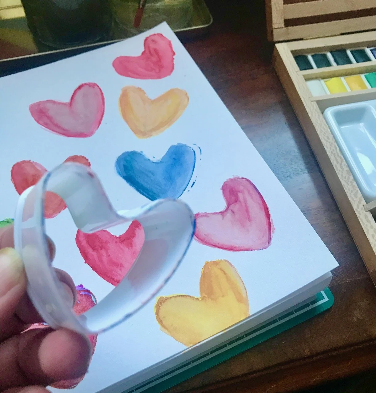 Holding a paint edged heart shaped cookie cutter above the sketchpad of painted hearts
