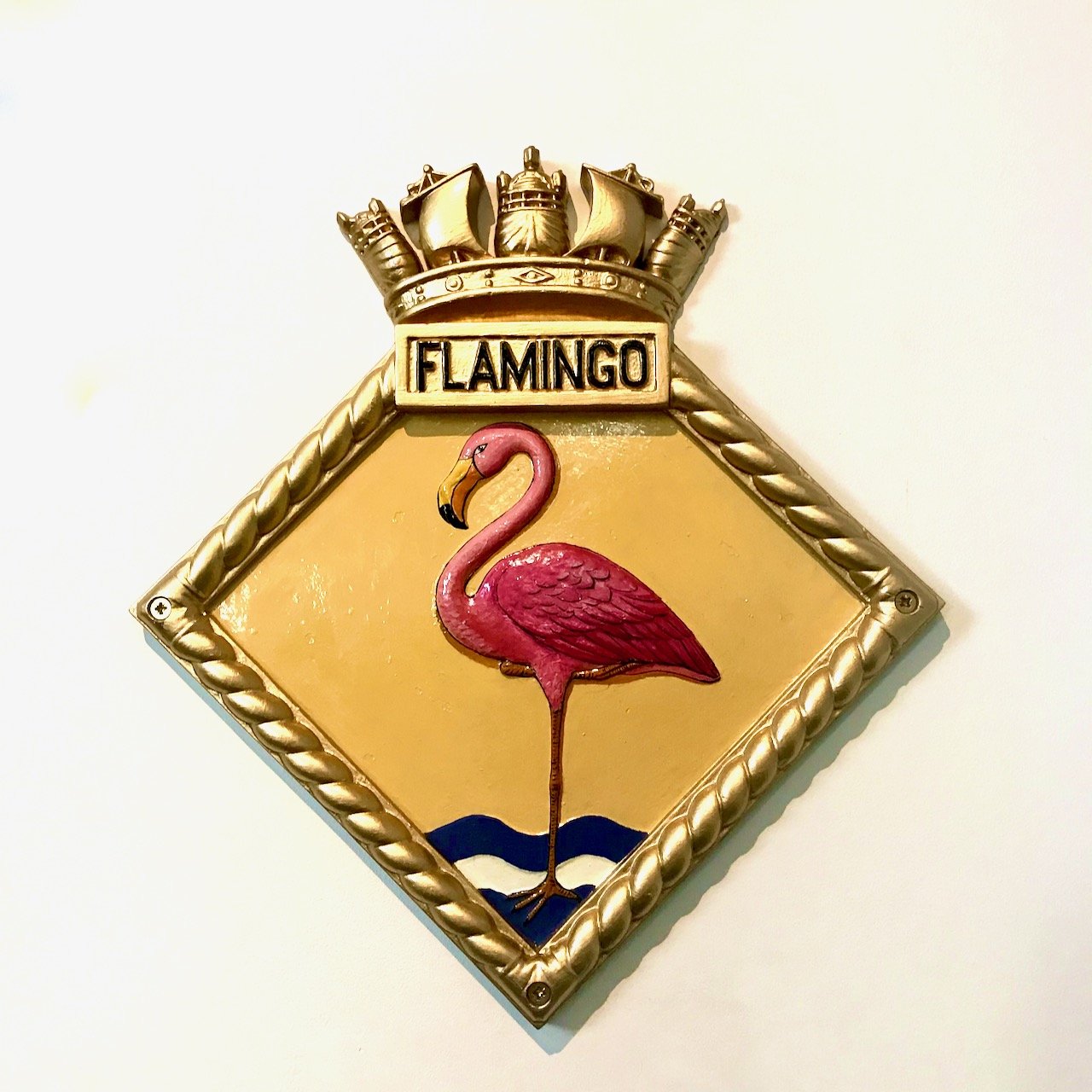 A ship's badge on the wall of the National Maritime Museum - diamond with a gold rope border, the background is yellow with a single pink flamingo in the centre, with the word flamingo above