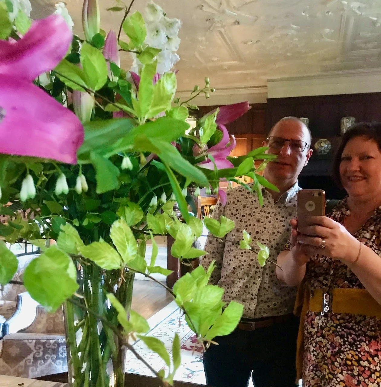 MOH & I in our finery photographing ourselves in a mirror sharing the frame with a large vase of green, white and pink flowers